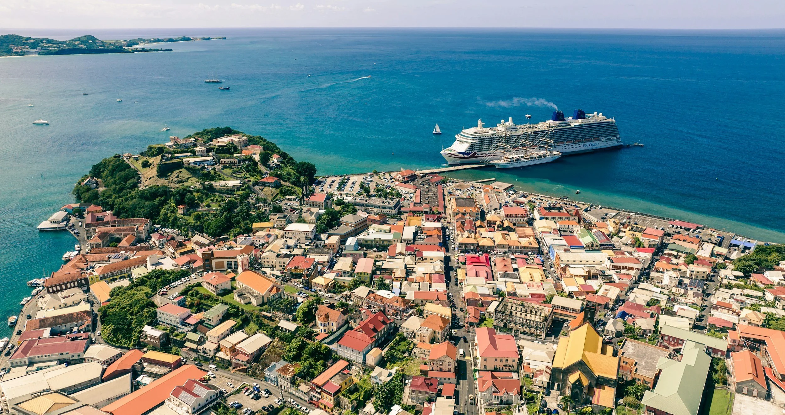 Aerial view of a coastal town with a large cruise ship docked at the harbor, surrounded by colorful buildings, greenery, and a few boats on the water.