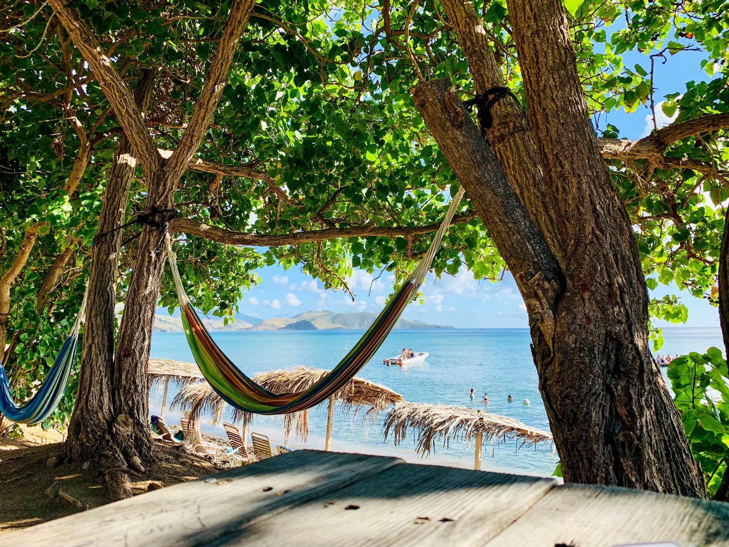 Beach scene viewed through trees, with a hammock hanging between the trees, a sandy shore with lounge chairs, a thatched umbrella, and a boat on calm blue water under a partly cloudy sky.