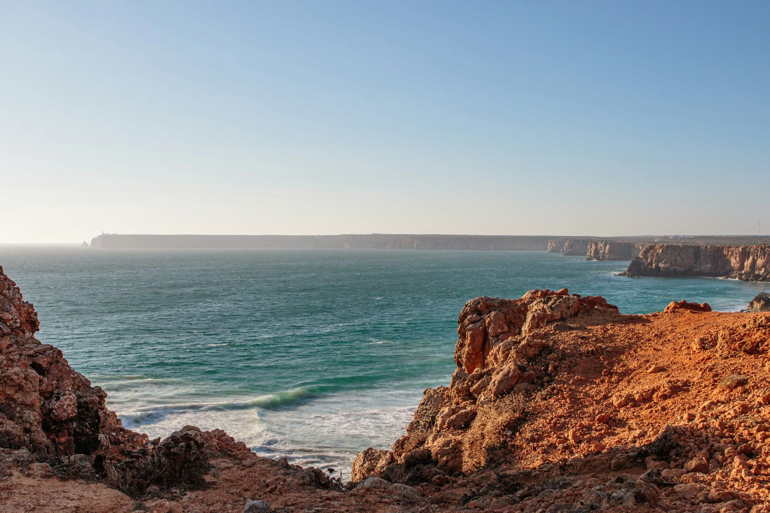 Cliffside view of the ocean with rocky cliffs in the foreground and a clear sky.