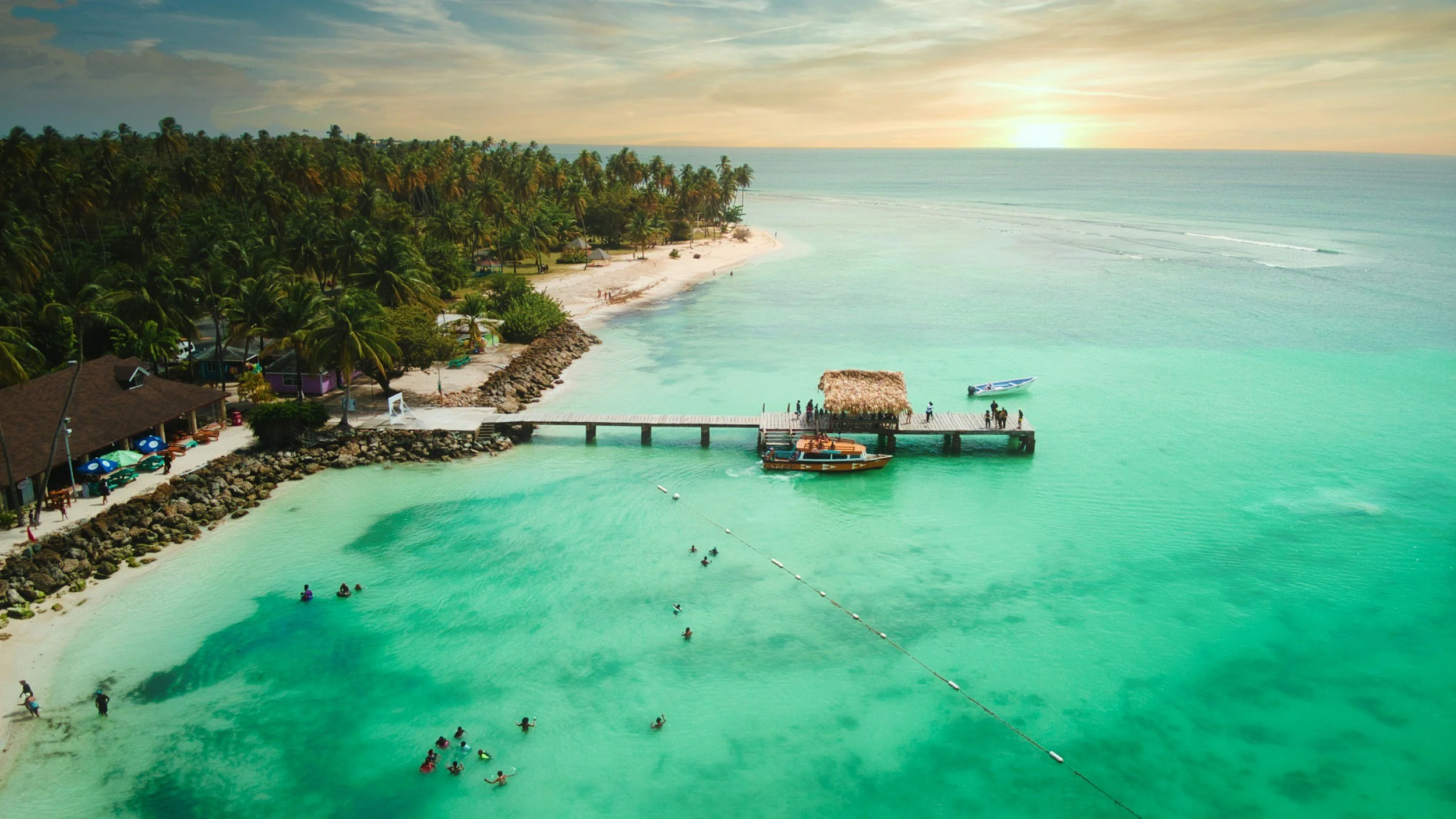 Tropical beach scene at sunset with palm trees, turquoise water, a pier with a thatched hut, boats, and swimmers in the water.