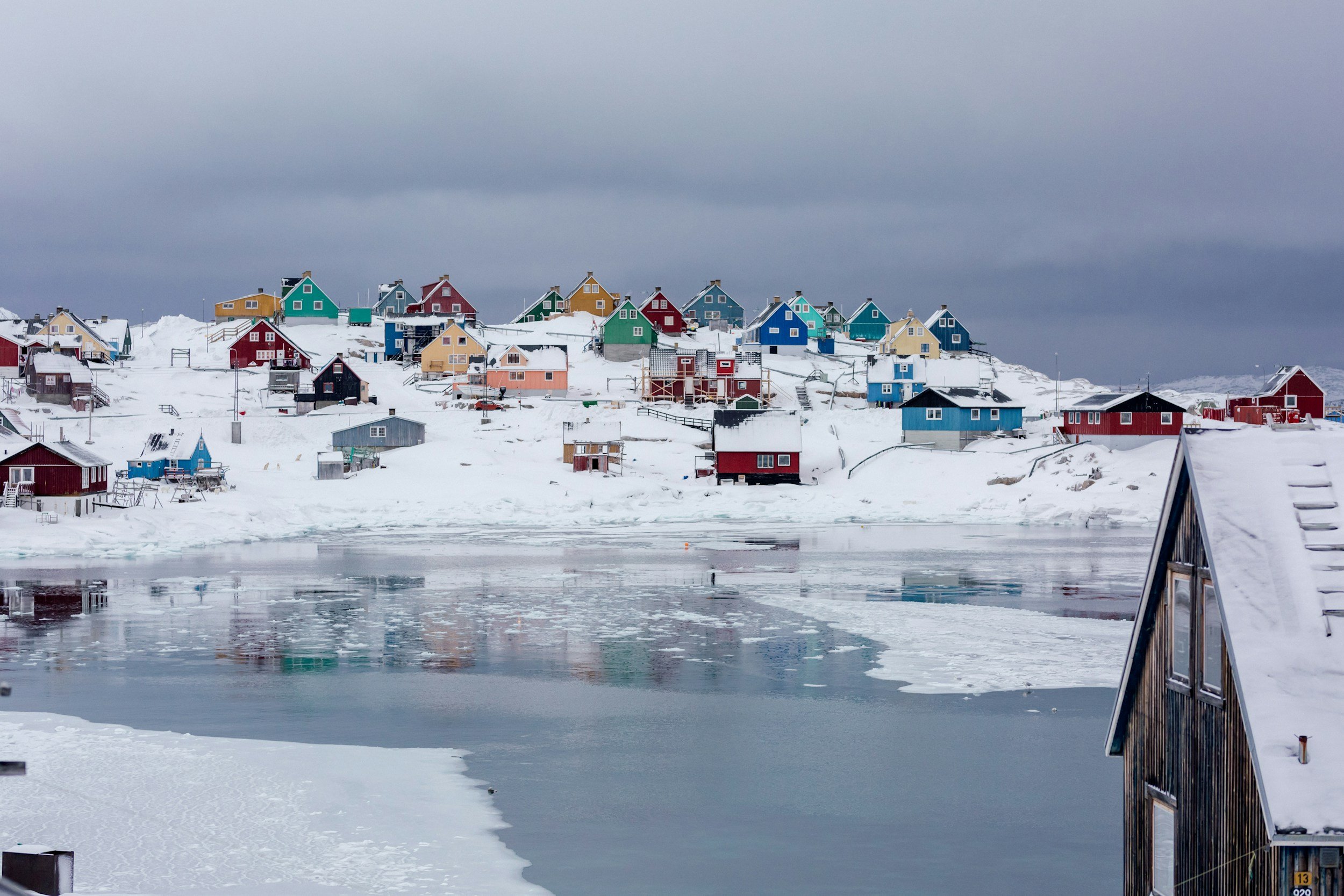 Colorful houses on a snow-covered hill by a partially frozen body of water under a gray, cloudy sky.