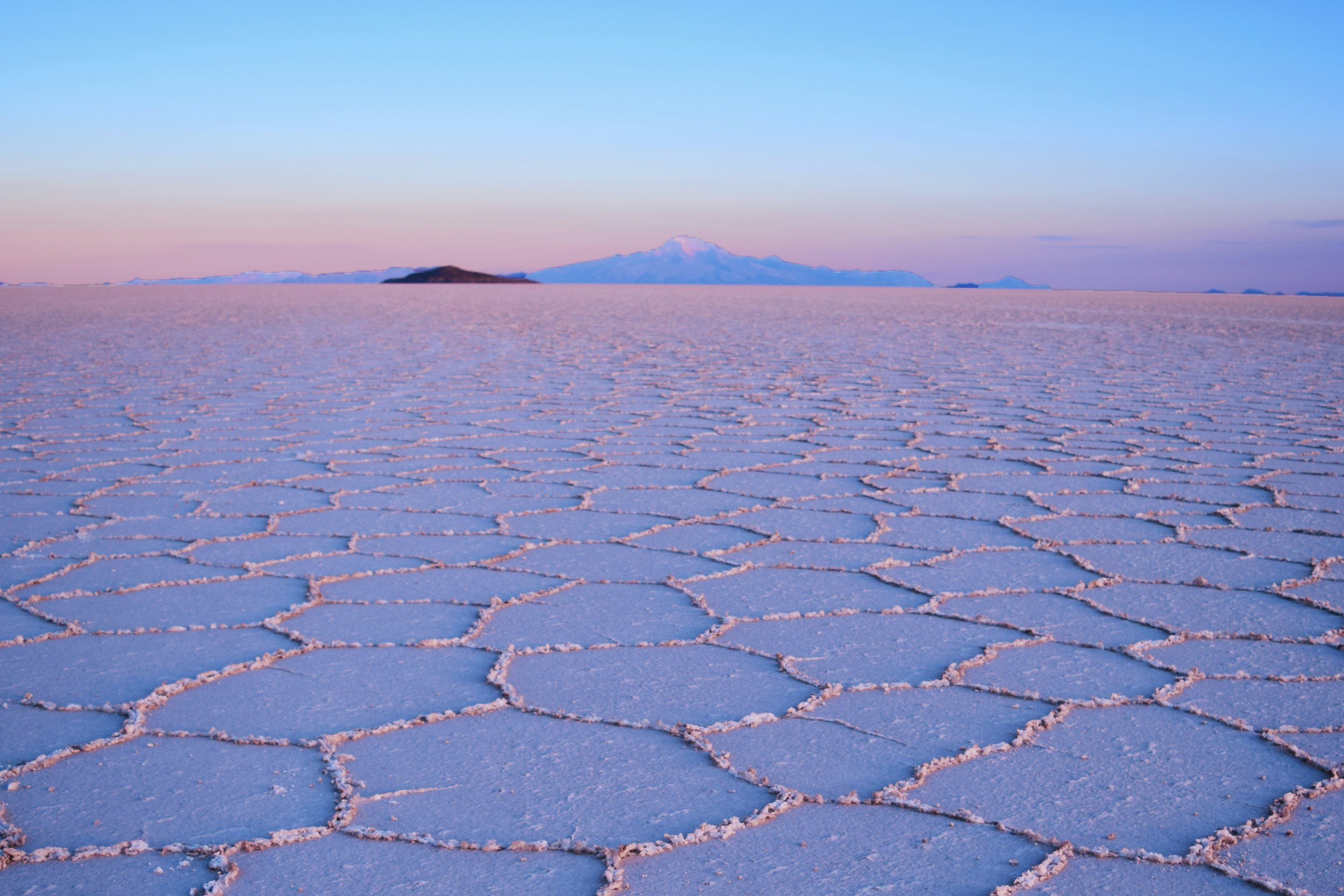 A salt flat with large hexagonal salt formations stretching to the horizon, with distant mountains under a clear sky.