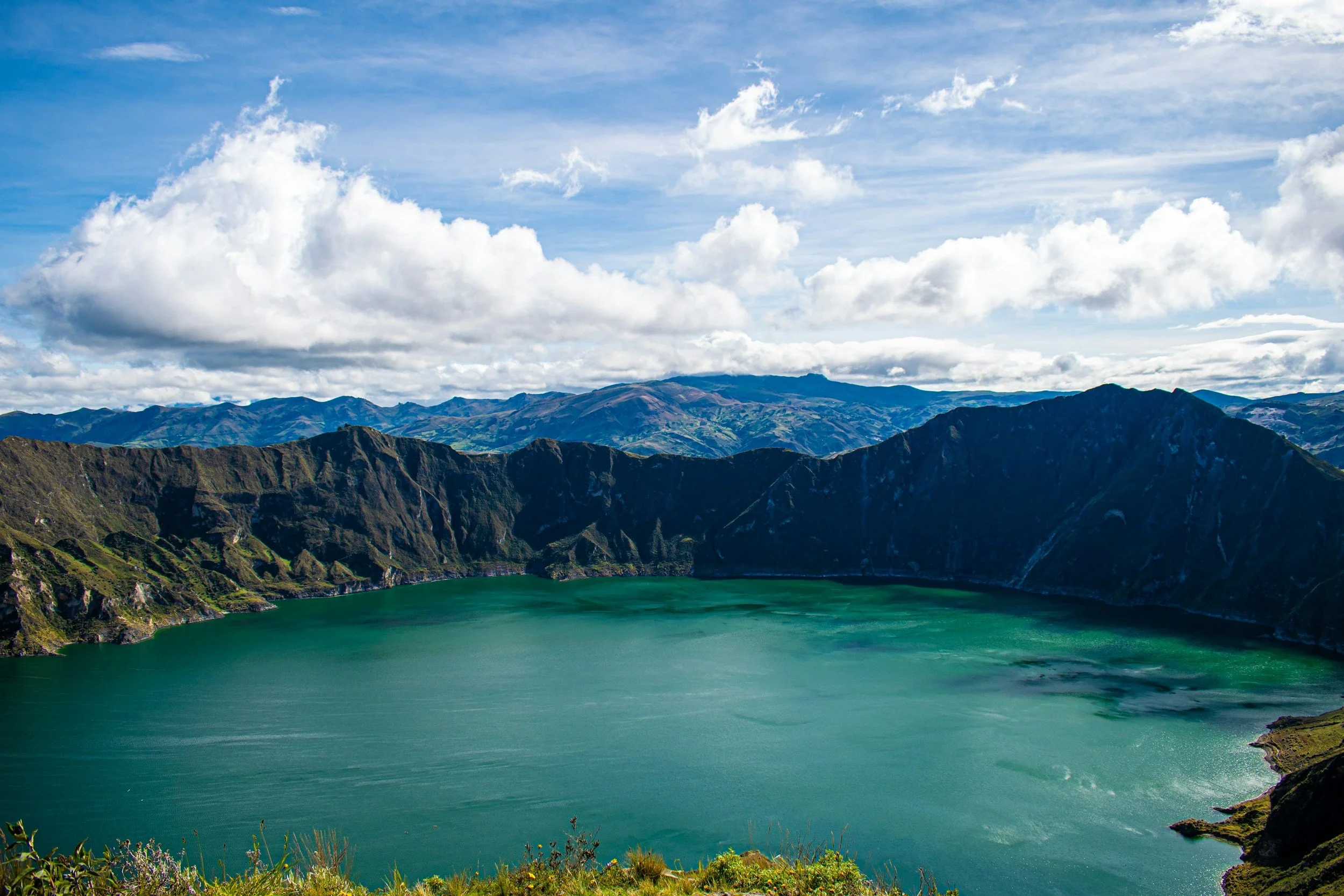 A crater lake surrounded by green mountains under a partly cloudy sky.
