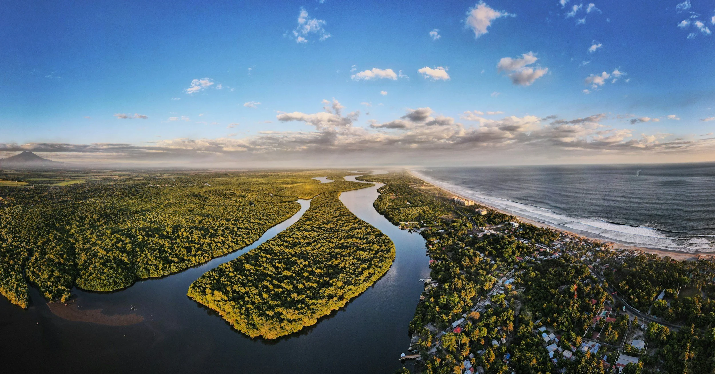An aerial view of a winding river flowing through a lush green forest near the coast, with a sandy beach and ocean waves on the right side, under a partly cloudy sky.