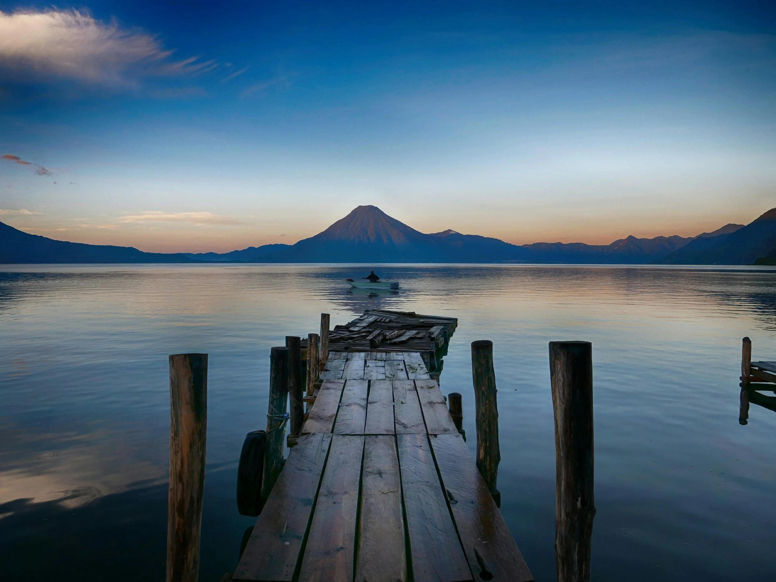 A wooden dock extends into a calm lake with mountains and a volcano in the background during sunset.