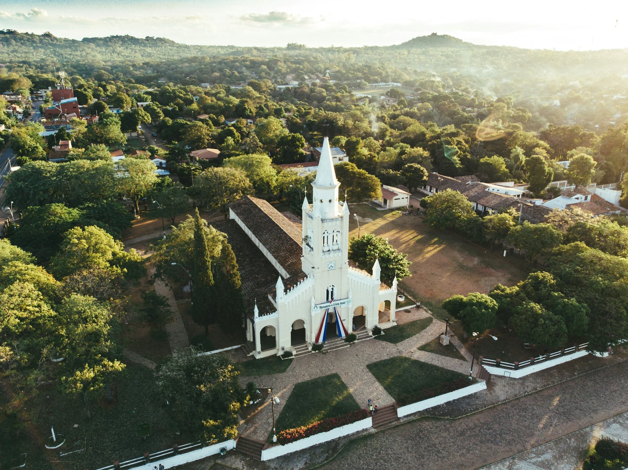 Aerial view of a white church with a tall steeple, surrounded by trees and a small fence in a green suburban area, with hills in the background and sunlight shining from the right.