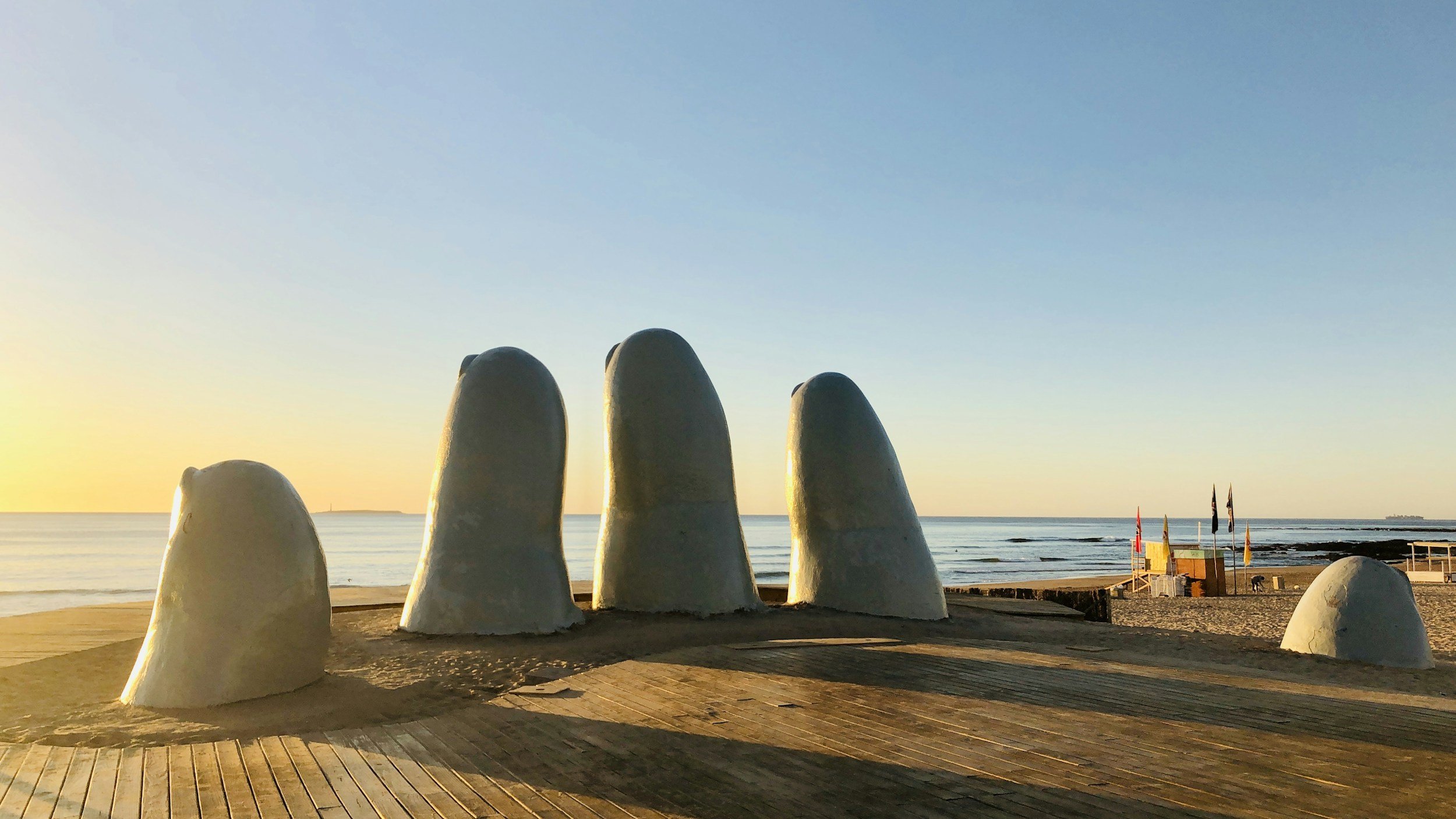 Beach scene with large white sculpture resembling five fingers or whale vertebrae, wooden pathway, sandy shore, calm ocean, and a clear sky at sunset.