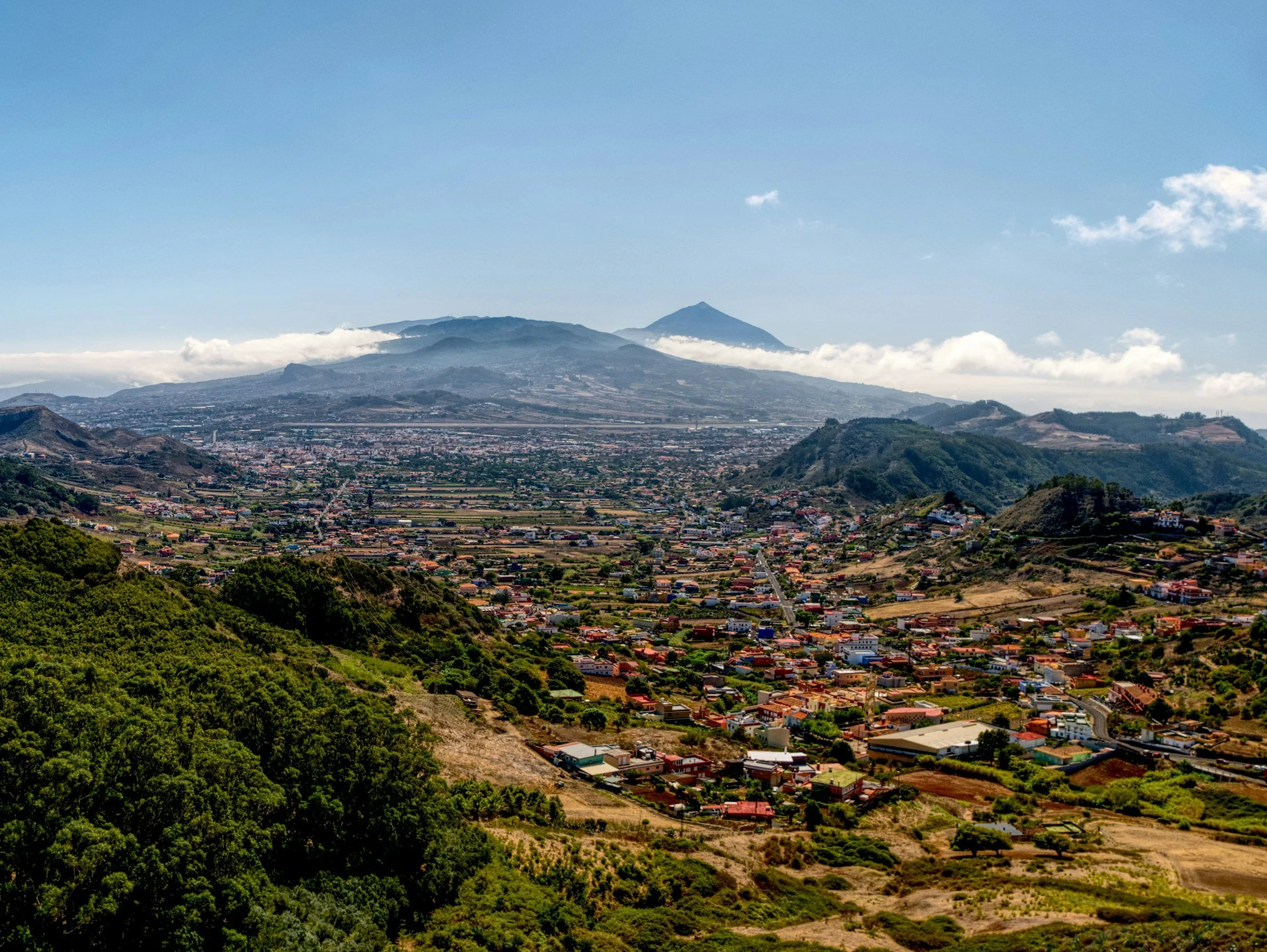 A scenic view of a town in a valley surrounded by green hills and mountains, with a volcano in the background under a partly cloudy sky.