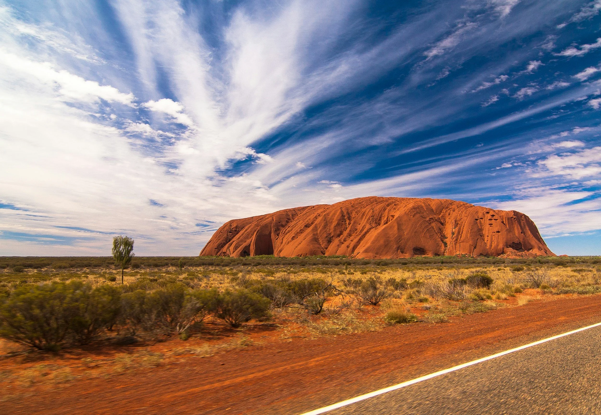 A red rock formation known as Uluru or Ayers Rock in the Australian Outback, with a partly cloudy sky overhead and desert vegetation in the foreground.