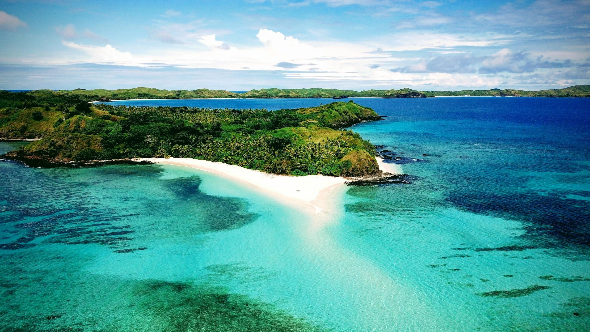 A tropical island with a white sandy beach, lush green vegetation, and turquoise waters, surrounded by the ocean under a partly cloudy sky.