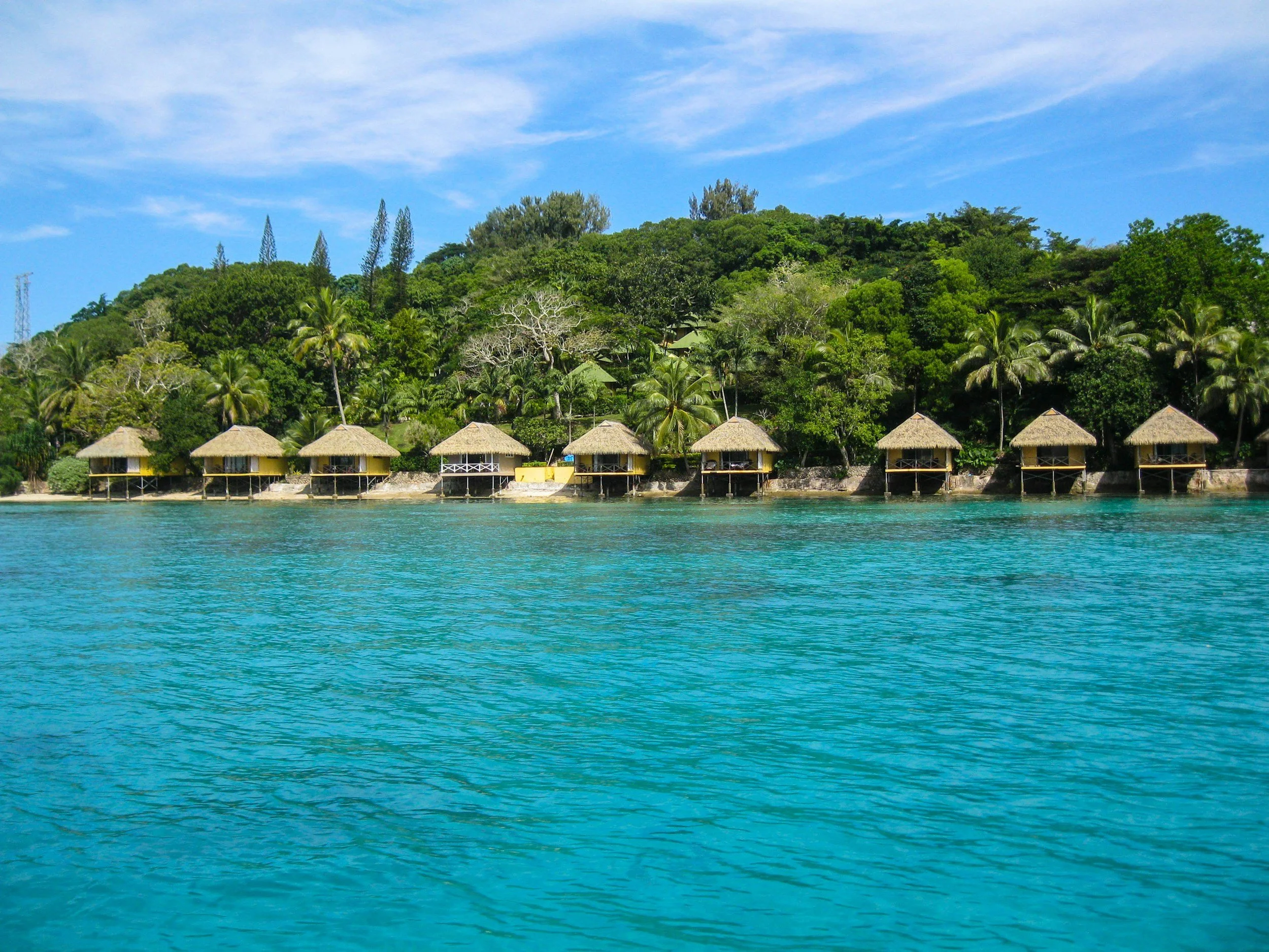 Beachfront of tropical island with seven thatched-roof huts on stilts over turquoise water, lush green trees and hills in the background, and a partly cloudy sky.