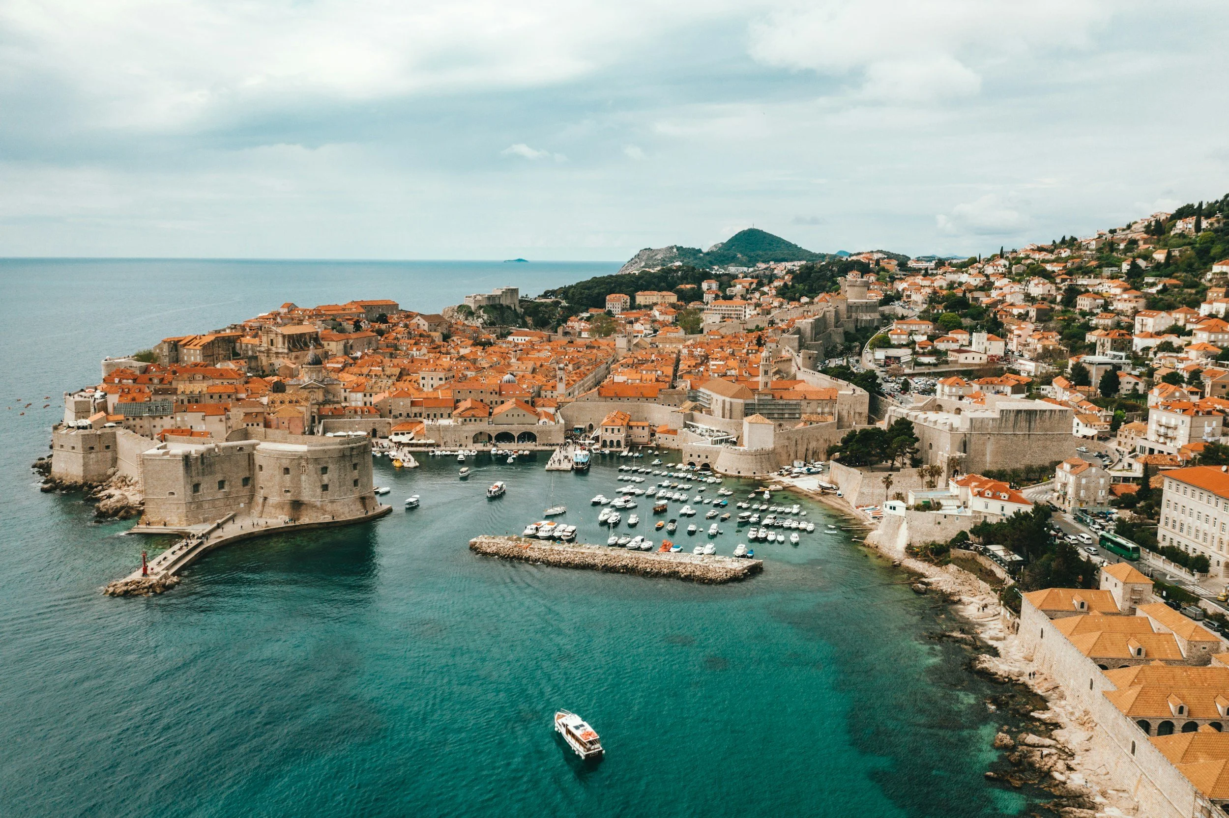 An aerial view of Dubrovnik, Croatia, showing historic stone walls, orange rooftops, and a marina with boats, overlooking the blue Adriatic Sea under a cloudy sky.