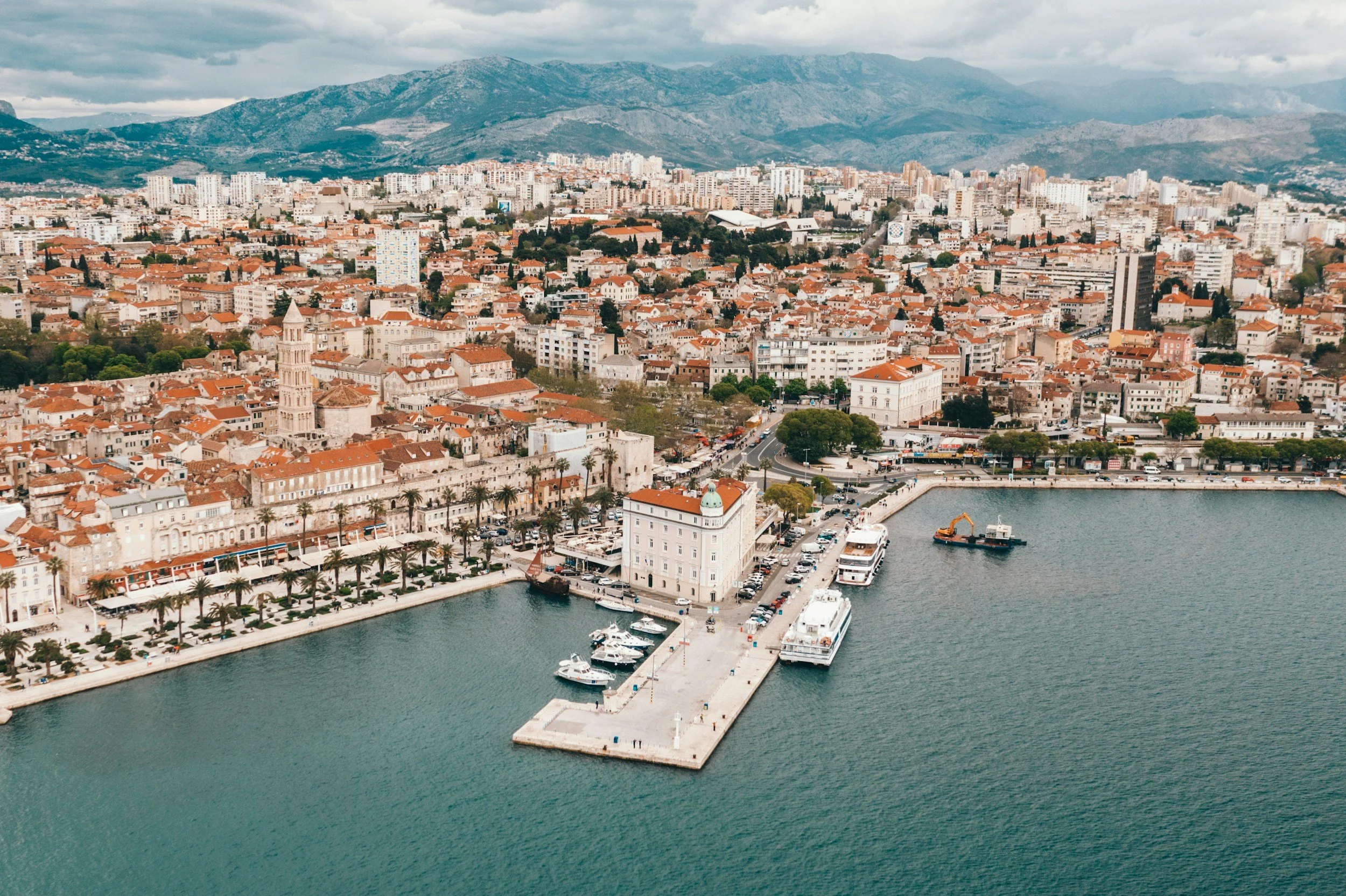 Aerial view of a coastal city with a harbor, boats, historic buildings with red-tiled roofs, a waterfront promenade lined with palm trees, and mountain landscape in the background.