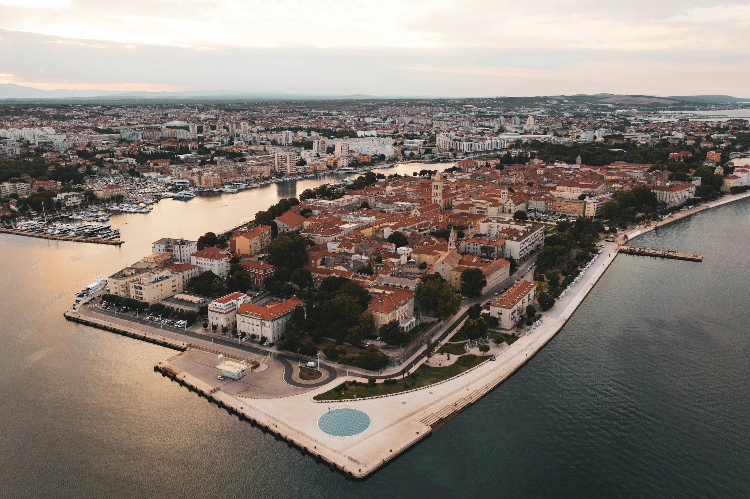 Aerial view of a historic town surrounded by water, with red-roofed buildings, a clock tower, greenery, and a waterfront promenade during sunset.