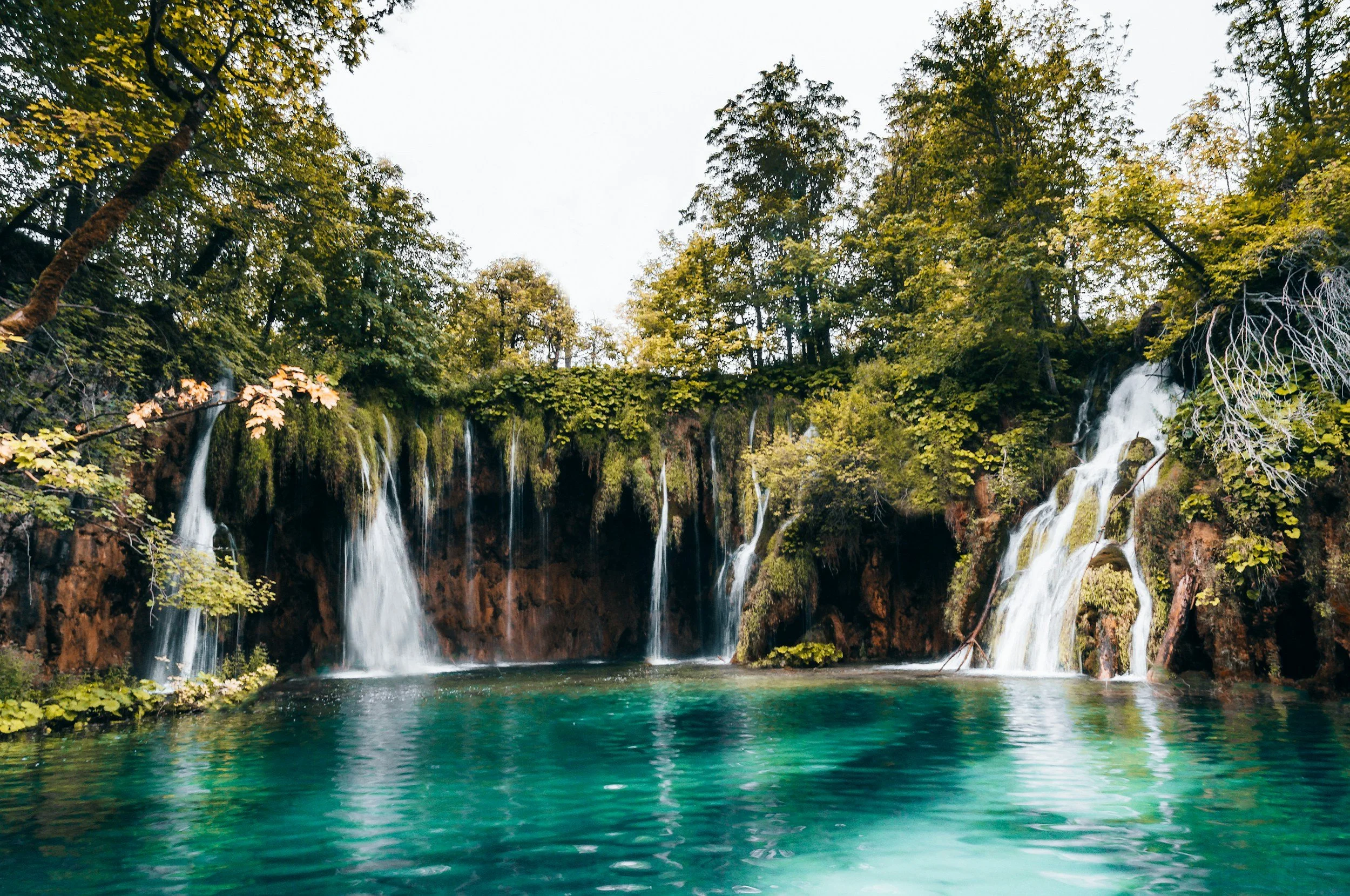 A scenic view of a waterfall flowing into a turquoise lake surrounded by lush green trees.