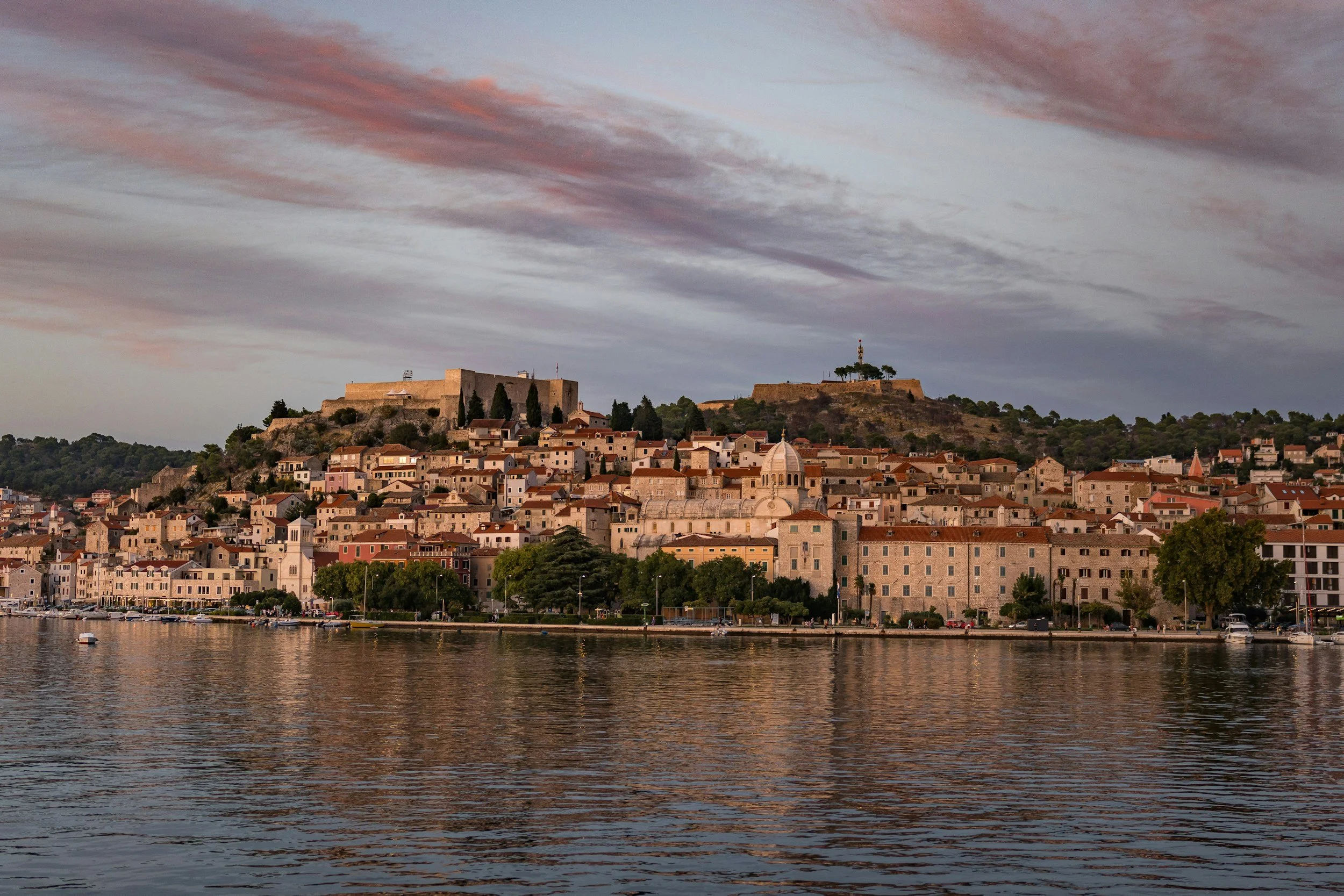 A coastal city with historic stone buildings and church towers, nestled on a hillside with fortresses on top, overlooking a body of water at sunset, with colorful clouds in the sky.