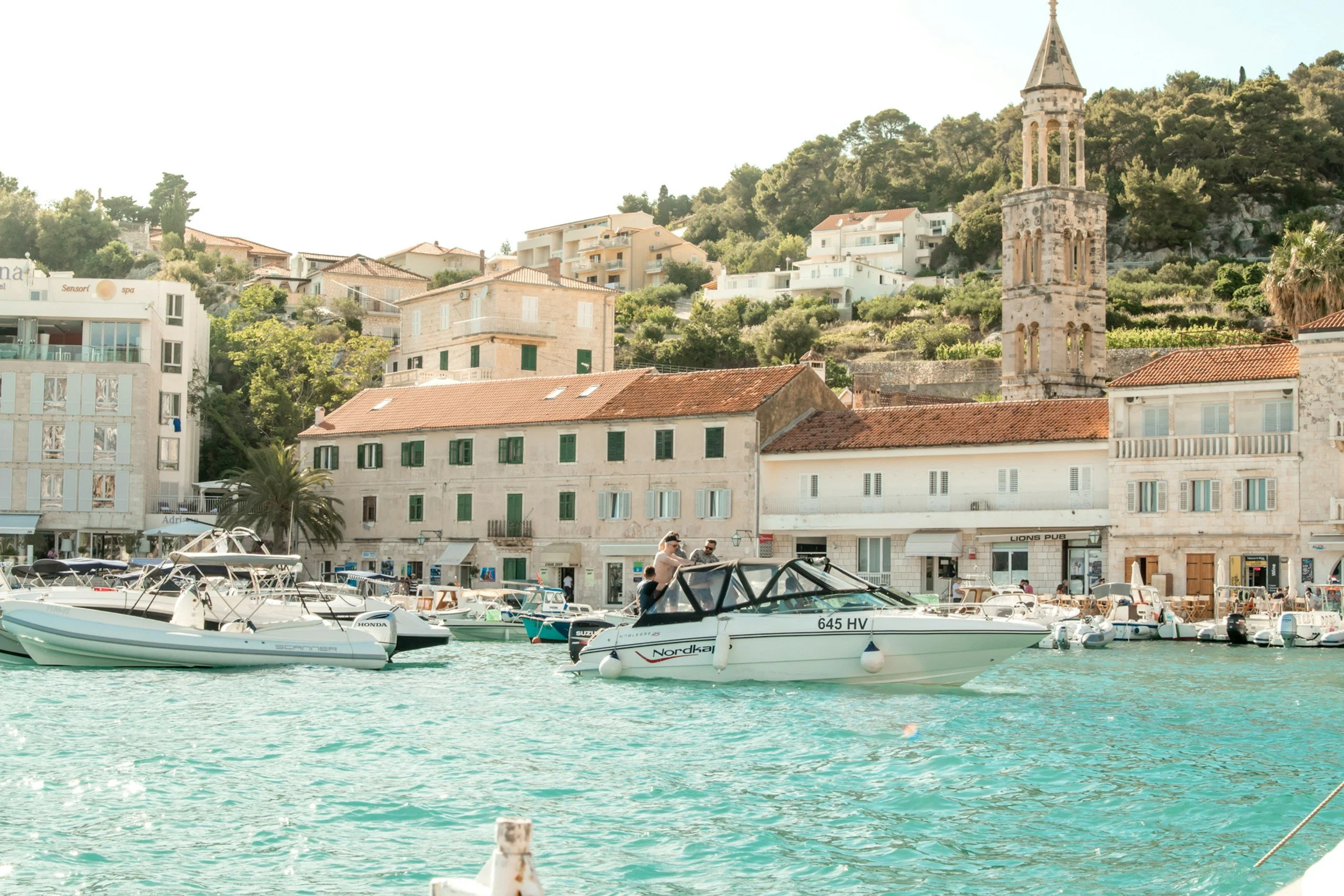 Boats docked in a harbor with a historic European town and church tower in the background, on a sunny day.