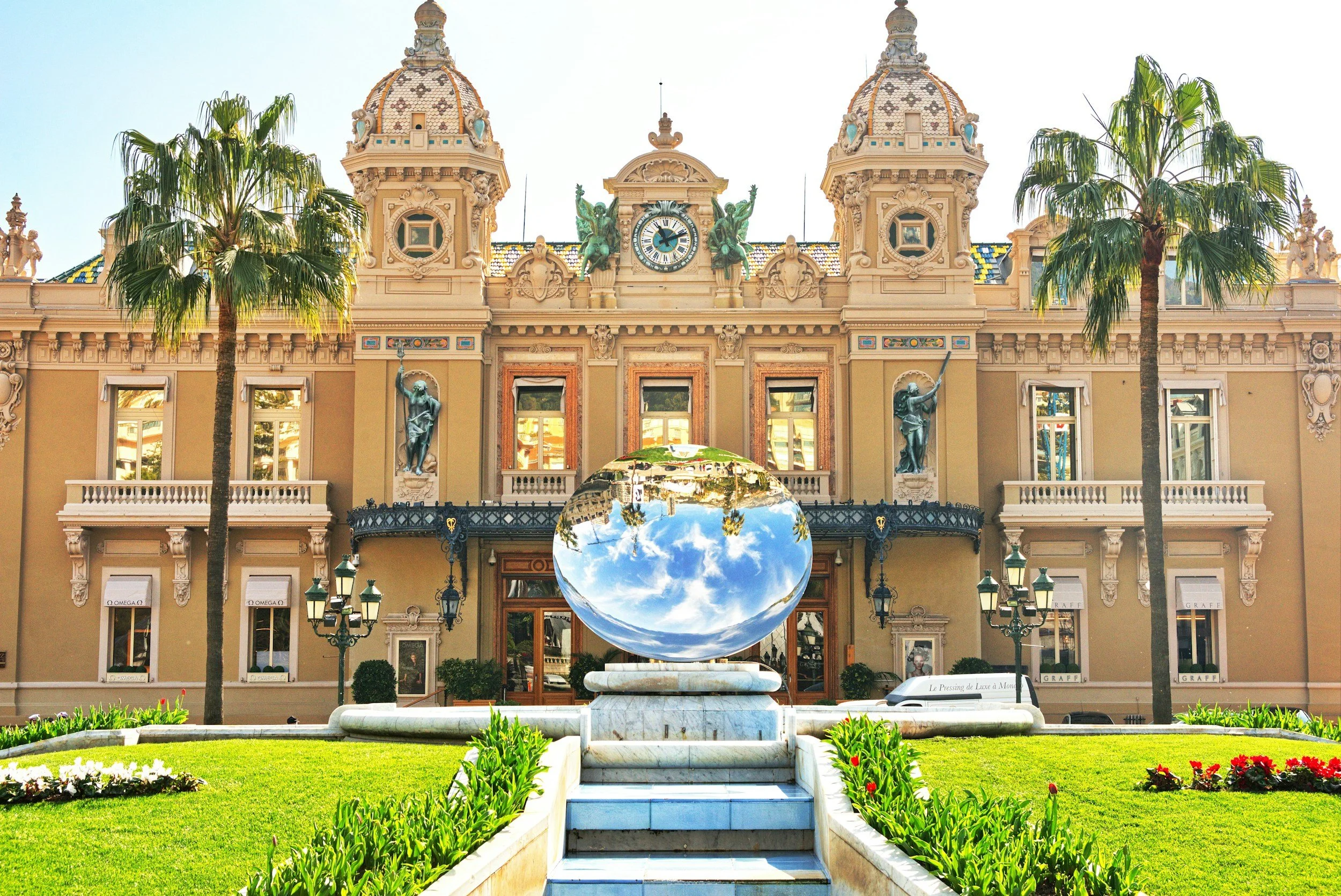 A grand ornate building with towers and statues in the background, landscaped gardens with green grass and colorful flowers in the foreground, and a large reflective spherical sculpture centered in front of the building that reflects the blue sky and clouds.