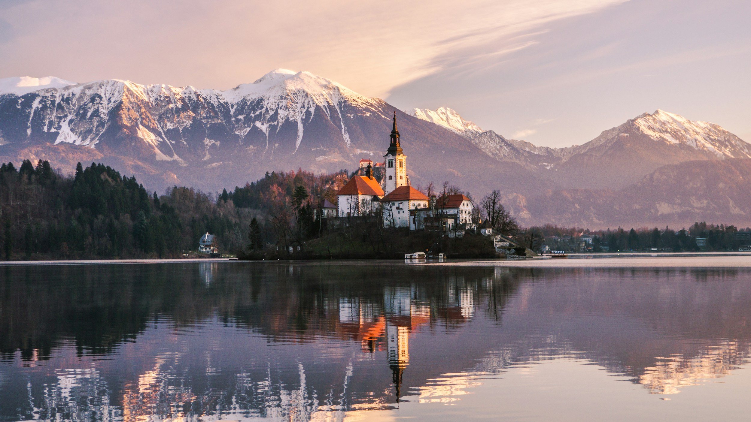 A church with a tall, pointed steeple and white walls located on a small island in a calm lake, surrounded by trees and mountains with snow-capped peaks in the background, reflecting in the water.