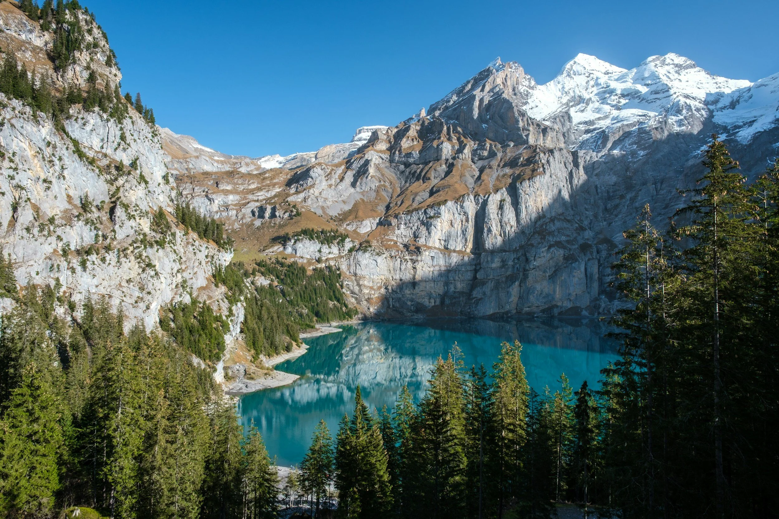 A mountain landscape with snow-capped peaks, steep rocky cliffs, a turquoise lake, and pine trees in the foreground under a clear blue sky.