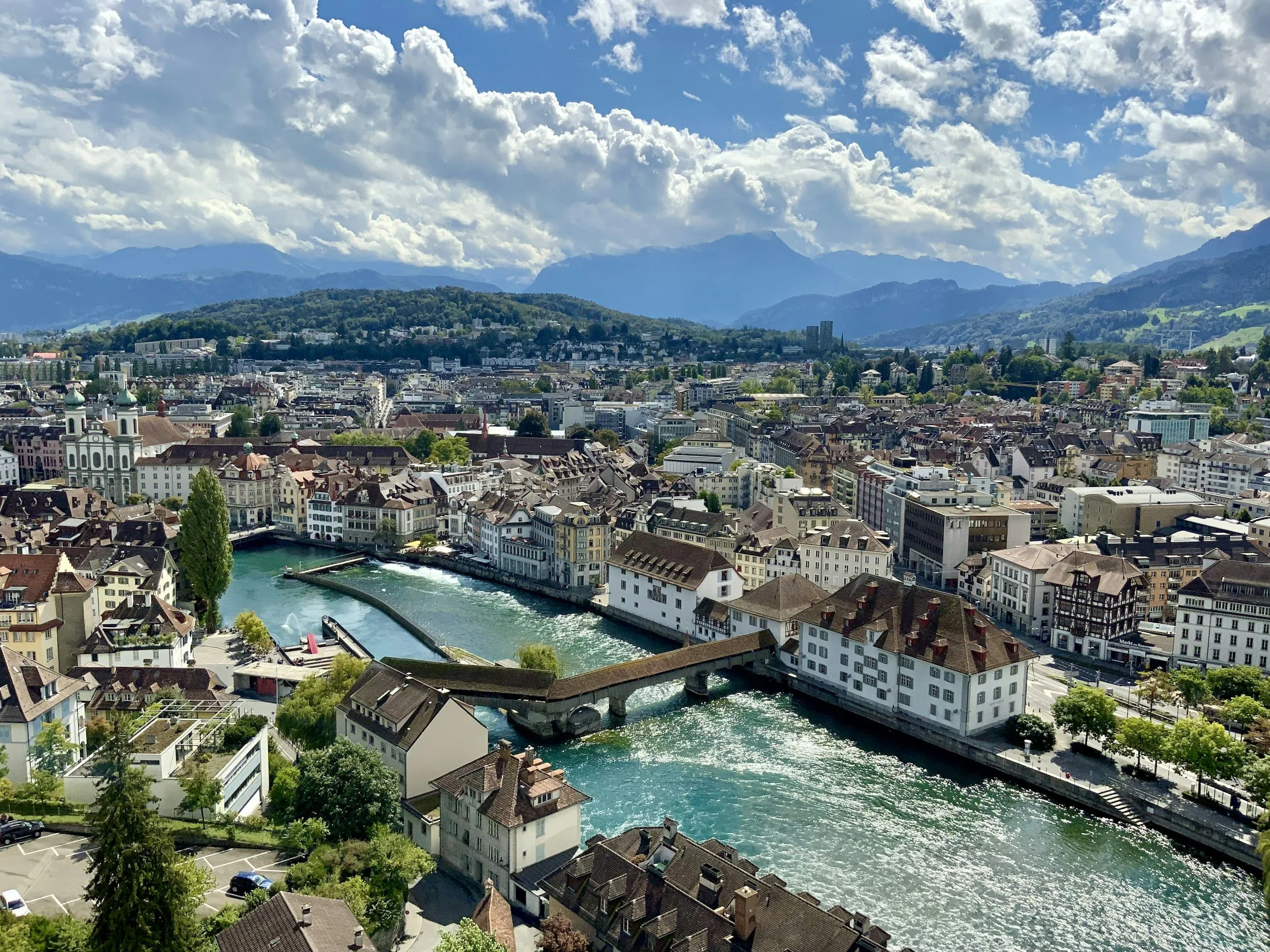 Aerial view of a city with a river running through it, surrounded by mountains and a partly cloudy sky.