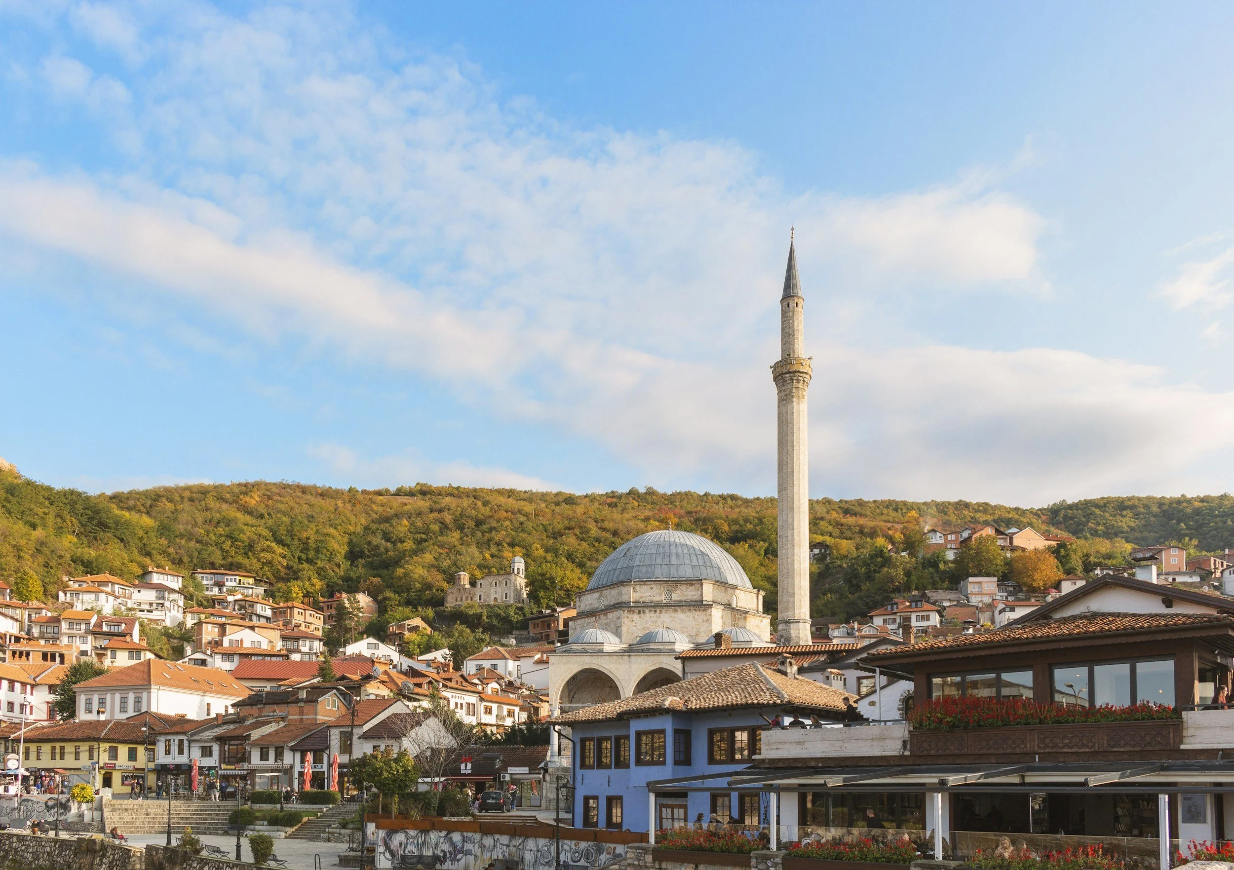 A cityscape featuring a mosque with a tall minaret, white and blue buildings, and a hillside covered with autumn trees under a partly cloudy sky.