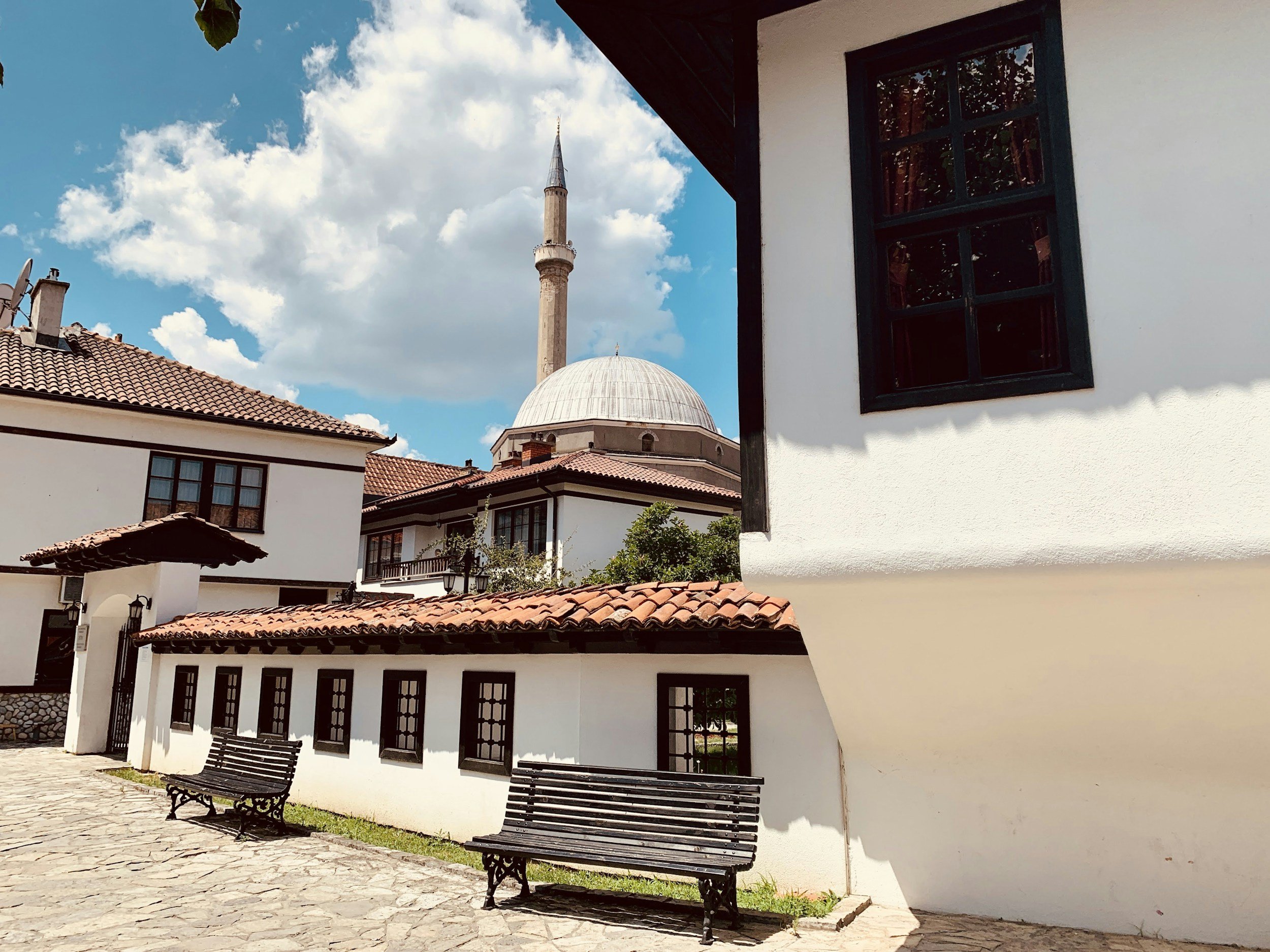 A street scene with white buildings featuring dark window frames and red-tiled roofs. In the background, there is a mosque with a dome and a tall minaret. The sky is bright with some clouds, and there are two black benches along a stone pathway in the foreground.