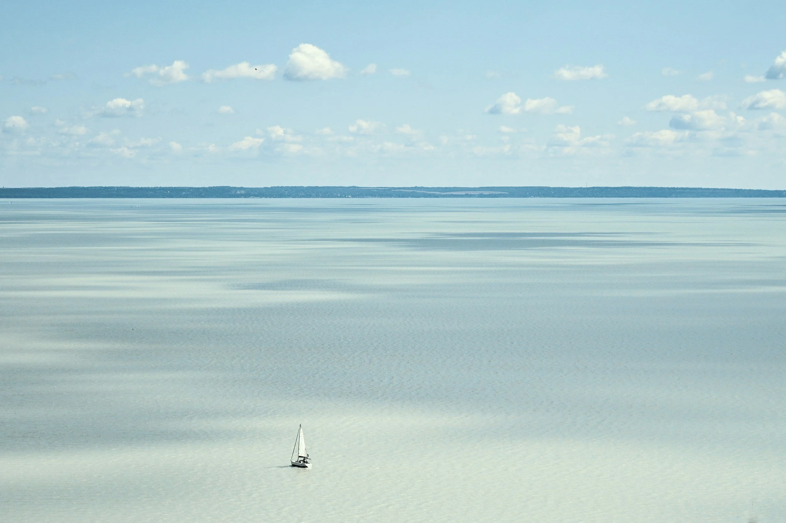 A sailboat floating on a calm body of water with a distant shoreline and a sky filled with scattered clouds.