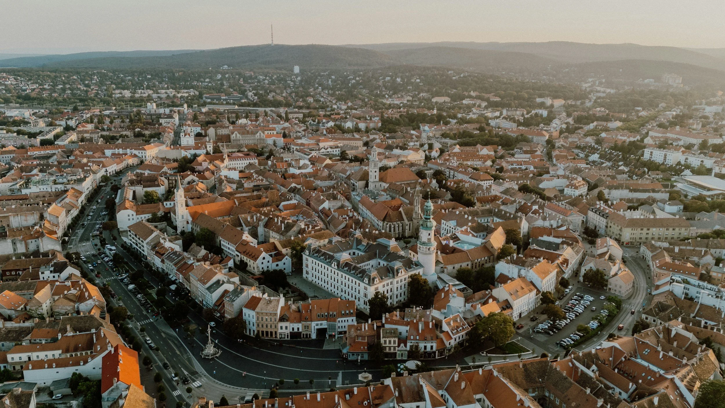 An aerial view of a historic city with red-tiled roofs, churches, and narrow streets, captured during sunset in Europe.