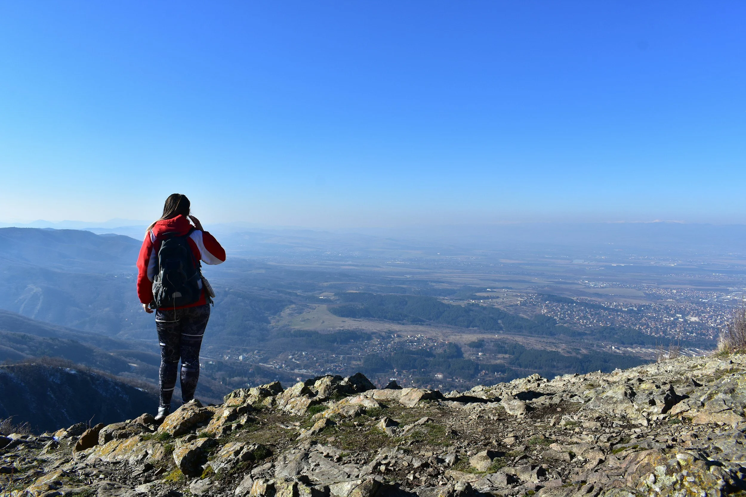 A person wearing a red and black jacket and patterned leggings standing on rocky terrain on a mountain, holding a phone and looking at the distant landscape of valleys, hills, and a city under a clear blue sky.