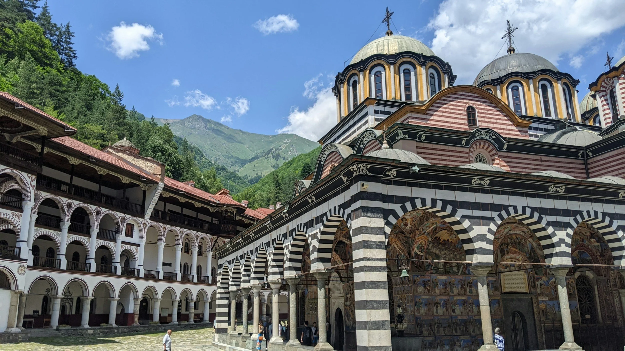 An ornate church with striped black and white walls, colorful frescoes, and domed roofs set against green mountains and a blue sky.