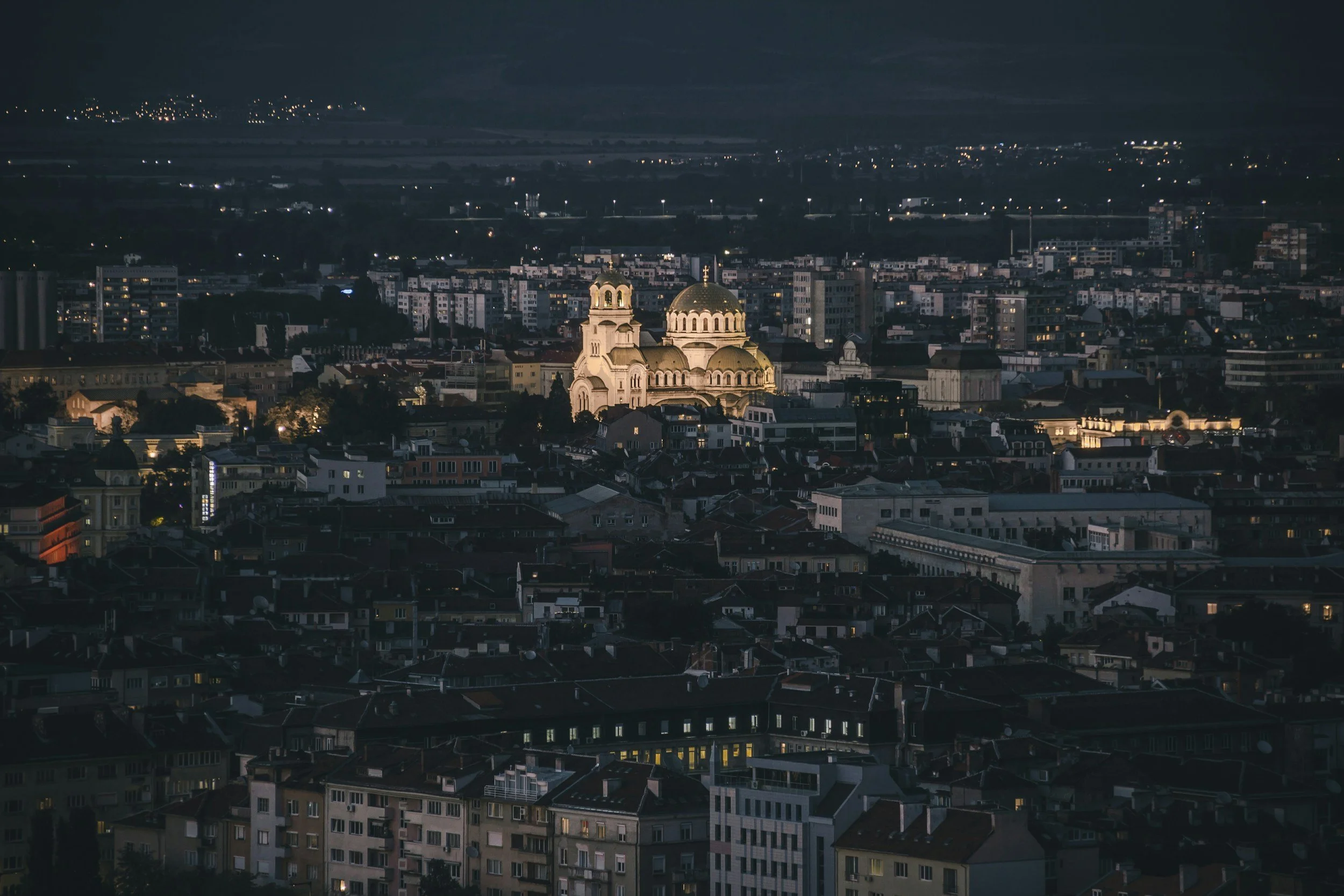 Nighttime cityscape with lit church in the center, surrounded by residential and commercial buildings, and distant hills in the background.