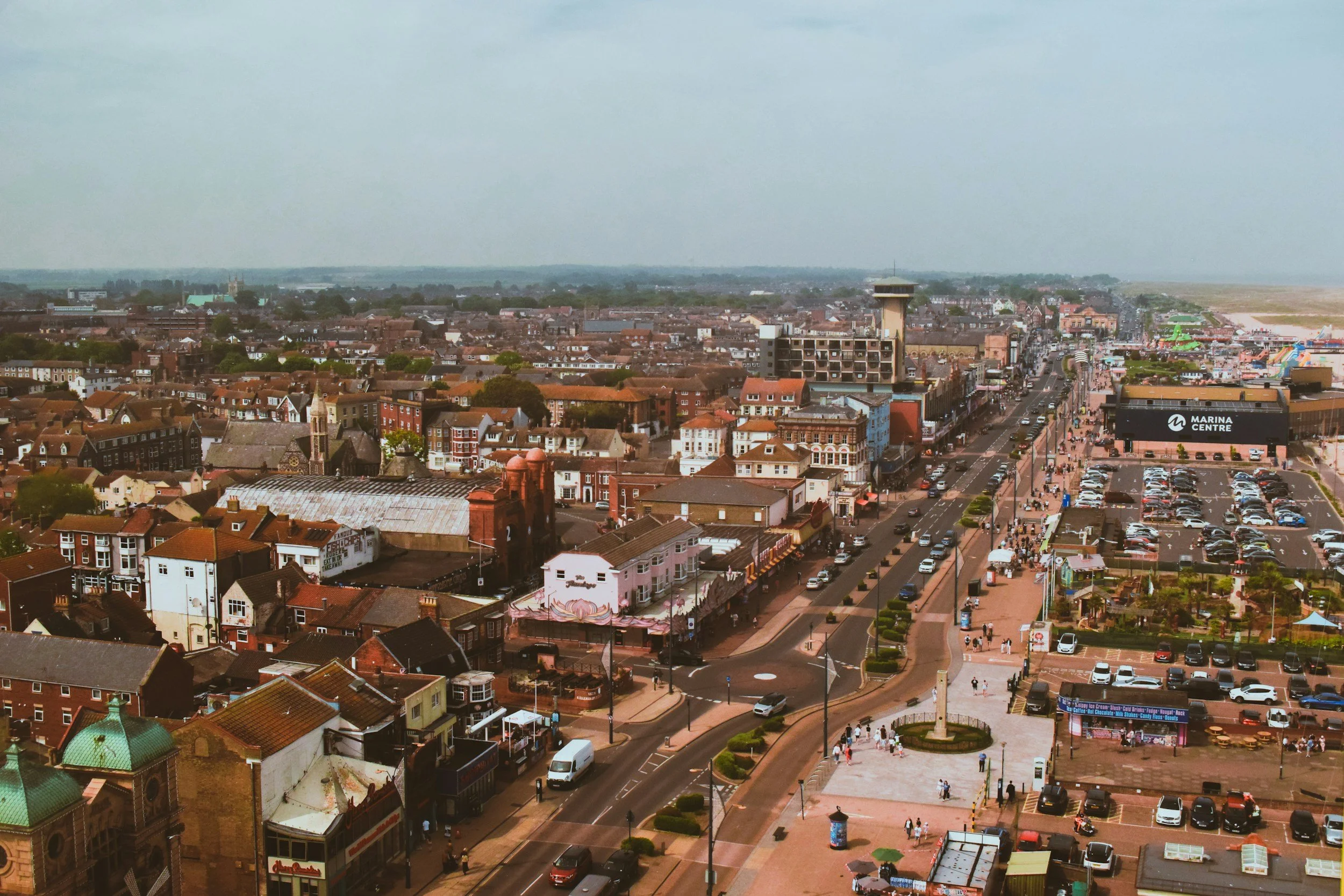 Aerial view of a busy town with a main street, parking lot, shops, and a wide promenade filled with people, with a distant waterfront and coastline under a partly cloudy sky.