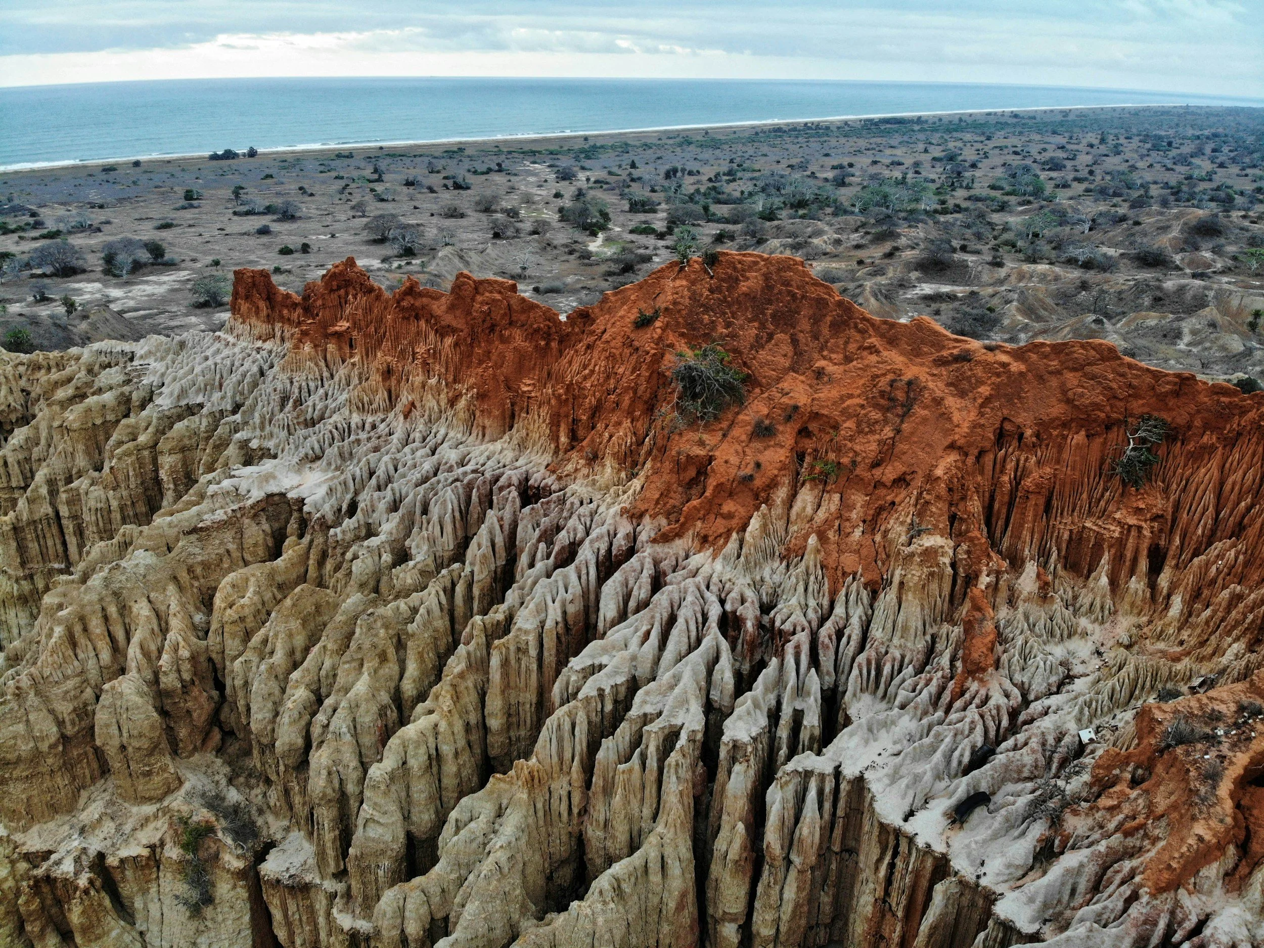 Aerial view of colorful layered rock formations near a sandy beach and expansive ocean under cloudy sky.
