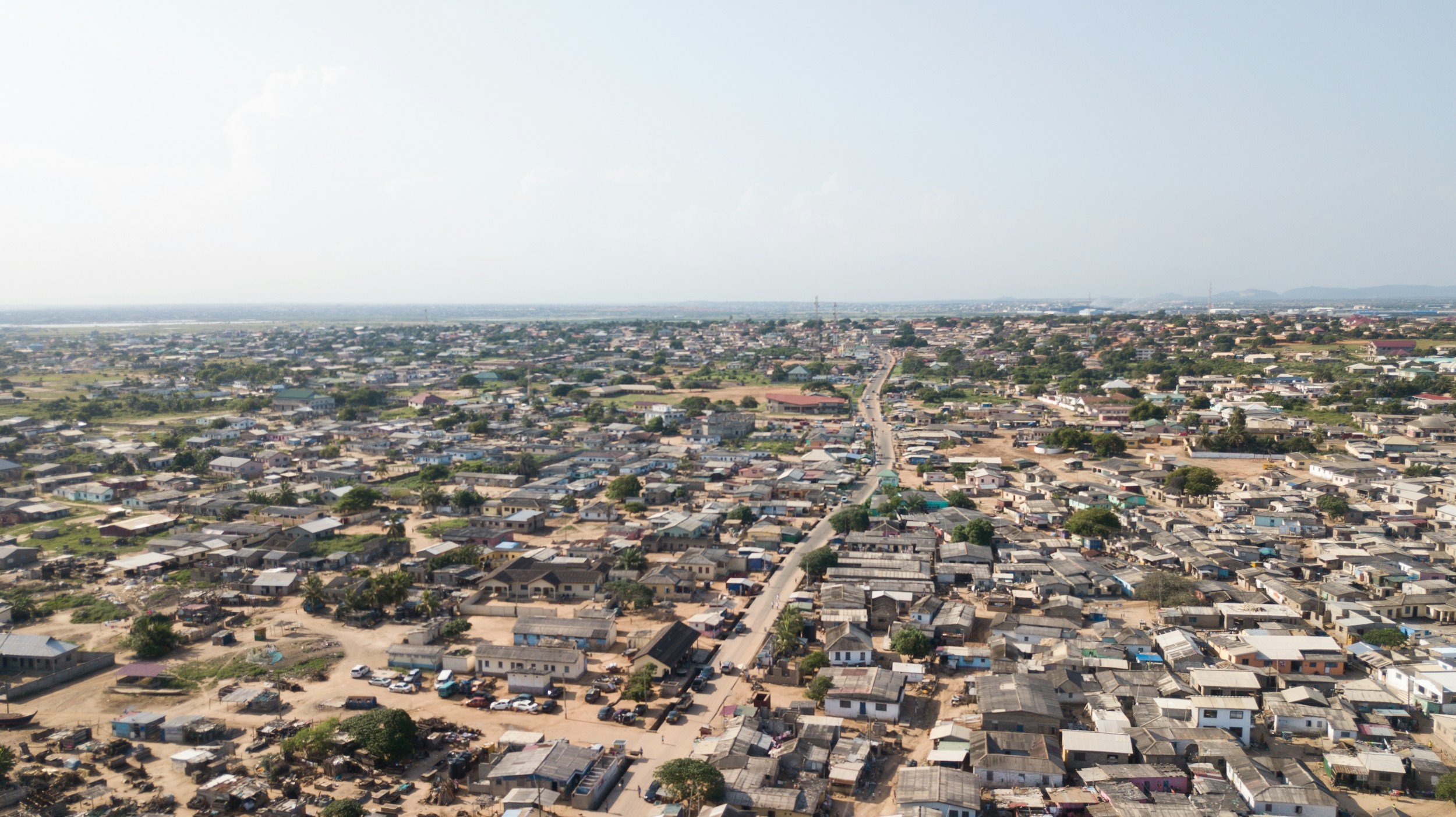Aerial view of a densely populated urban area with small houses and buildings, a main road running through the neighborhood, and open land in the distance.