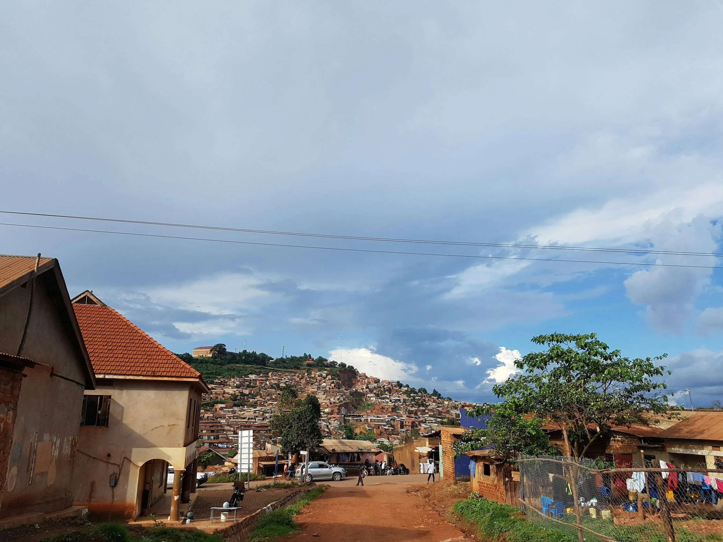A view of a hillside town with numerous houses, some trees, and a partly cloudy sky. In the foreground, there are a few houses, a dirt road, and some parked cars and people walking.