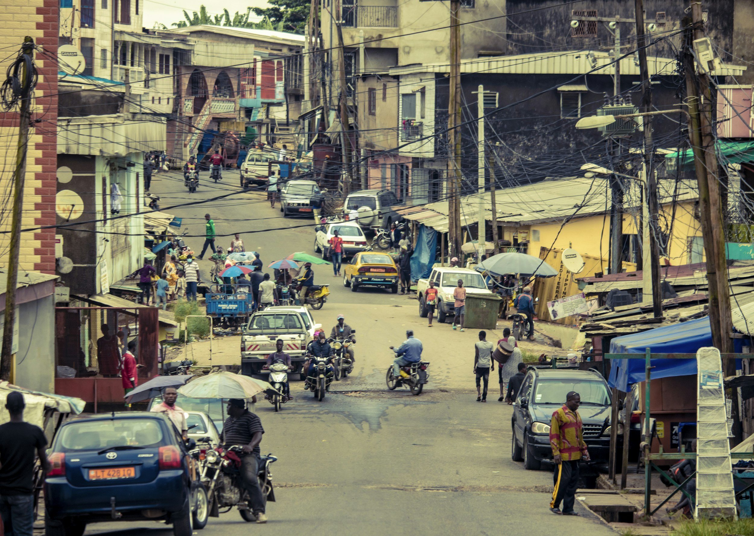 A busy urban street scene with cars, motorcycles, and pedestrians, small market stalls, and buildings with overhead power lines.