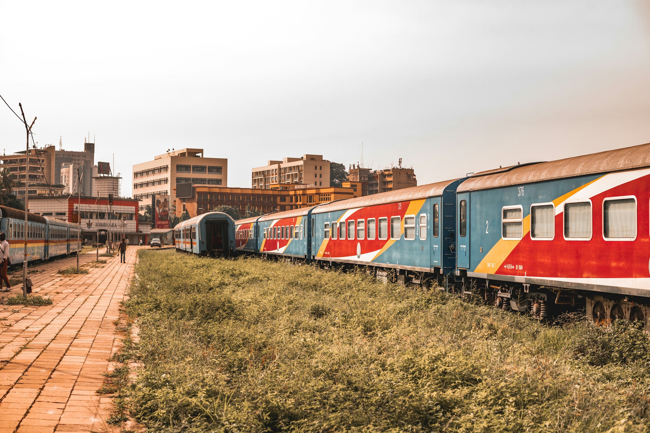 Colorful train parked on tracks in an urban area with buildings and a walkway nearby.