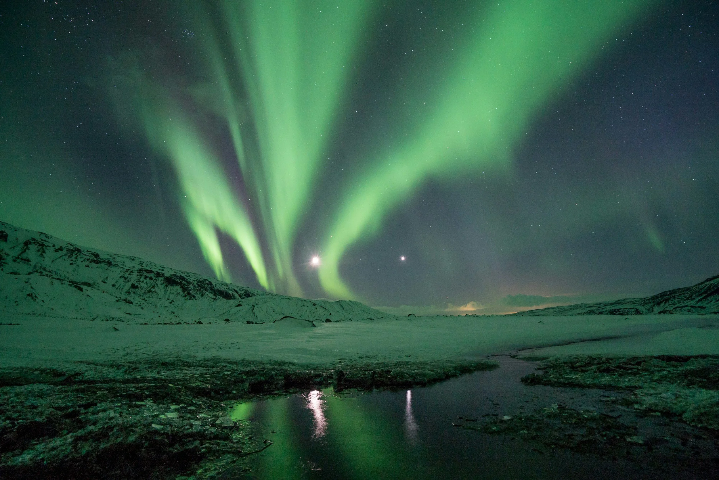 Northern lights illuminating the night sky over a snowy landscape with a small body of water in the foreground.