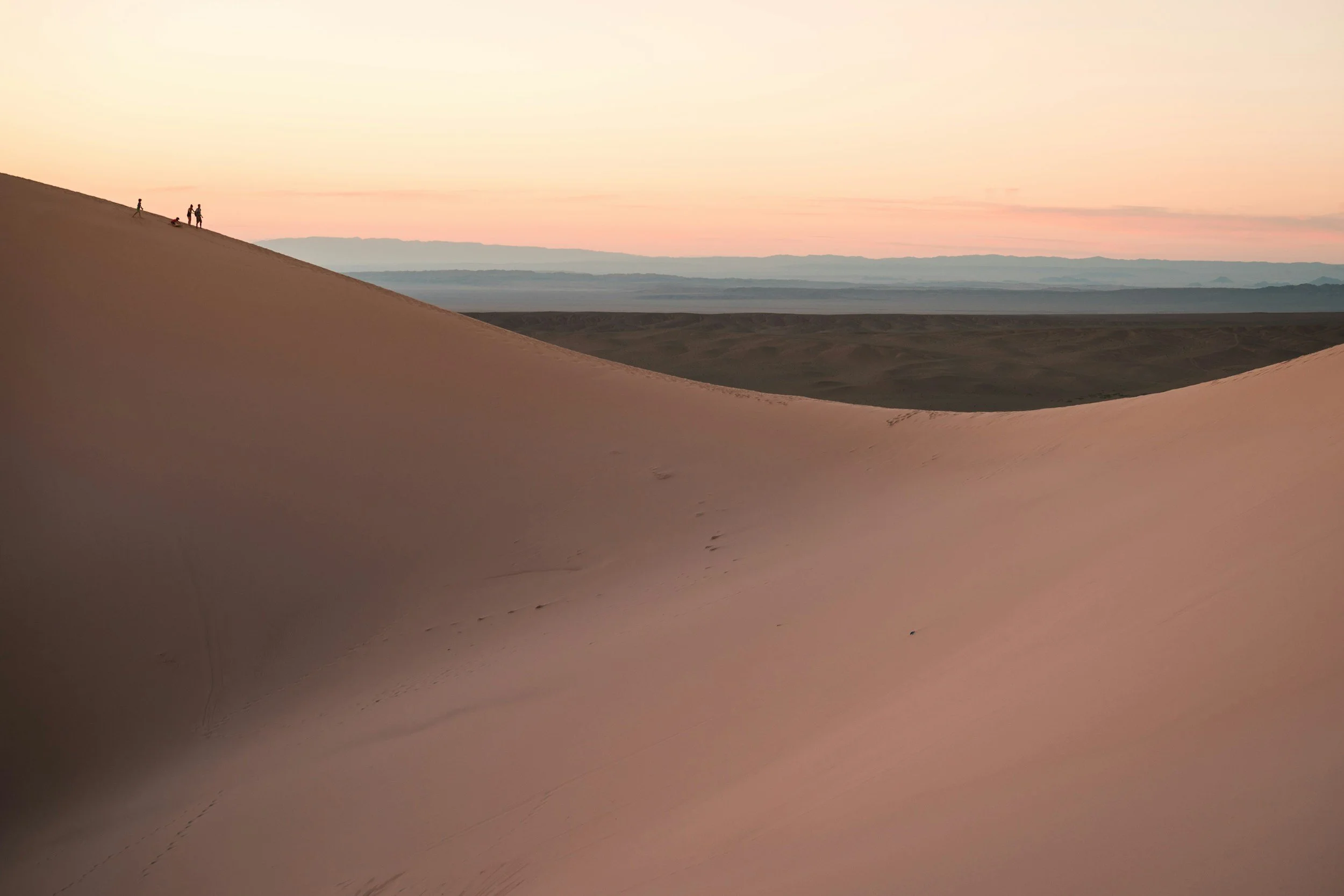 A desert landscape during sunset with rolling sand dunes and a few people walking on the crest of a dune in the distance.