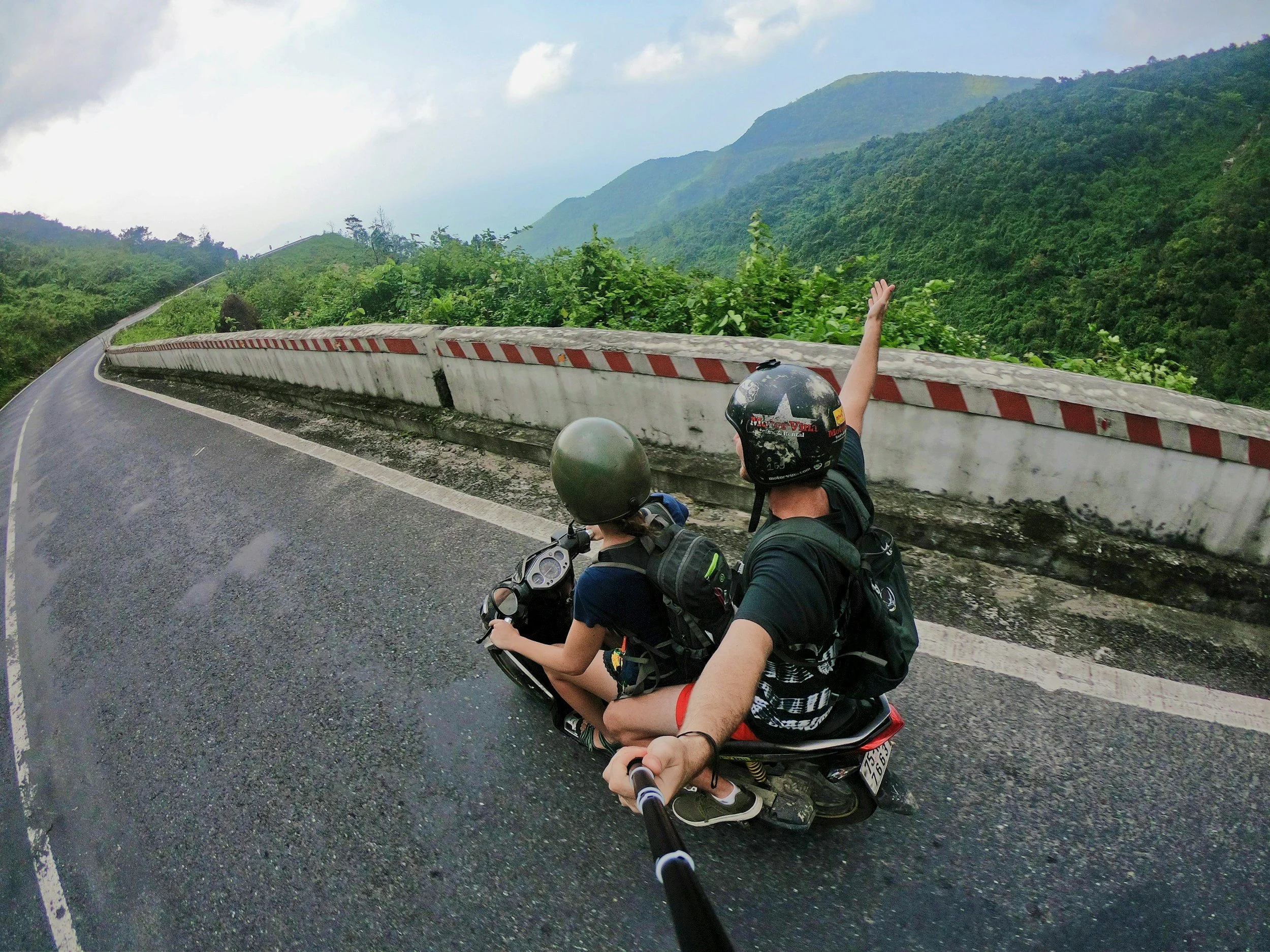 Two people riding a scooter with a child passenger, all wearing helmets, on a winding mountain road with lush green hills in the background.