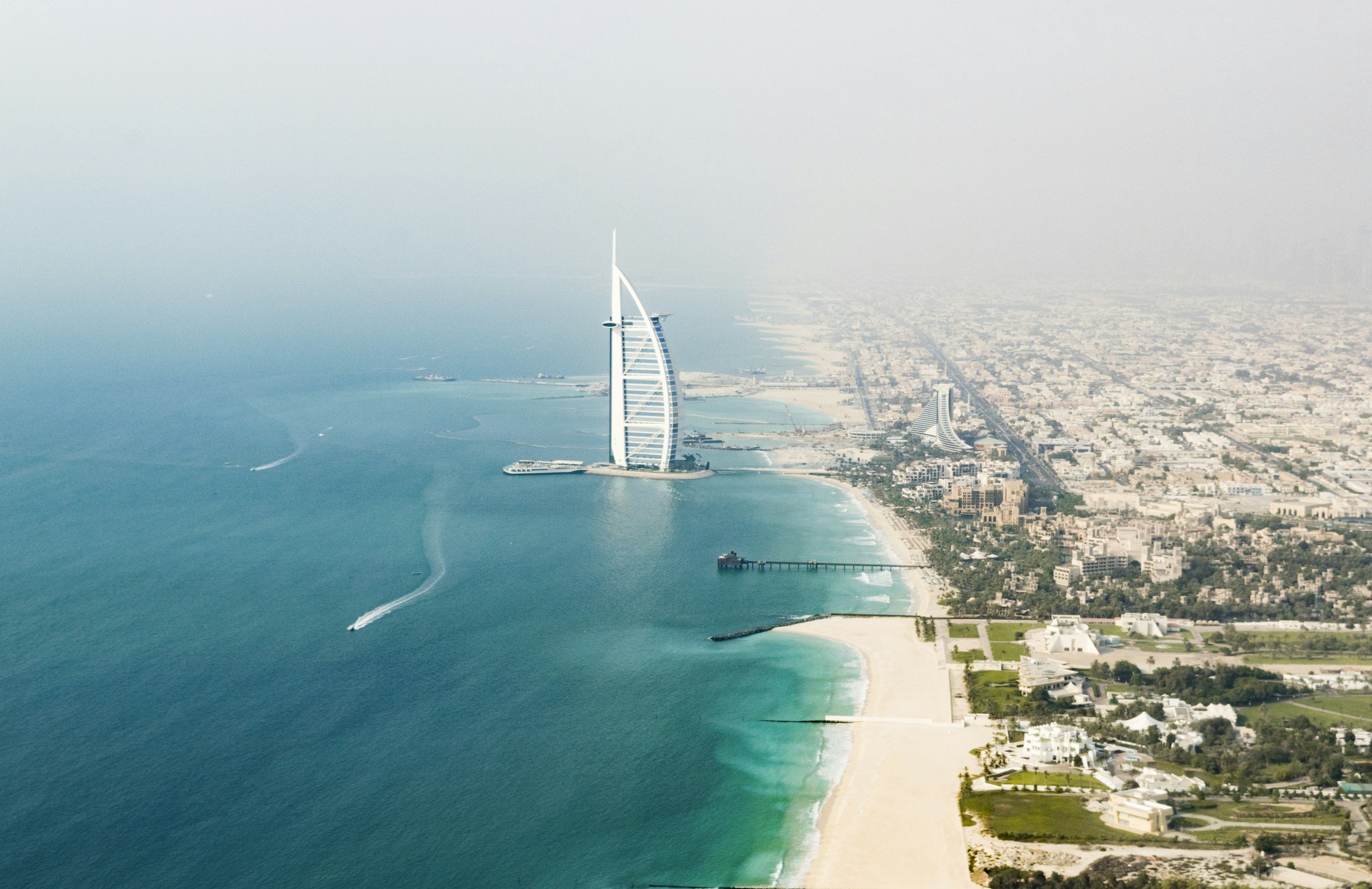 An aerial view of Dubai coastline featuring the Burj Al Arab hotel, sandy beaches, and a city with numerous buildings extending inland.