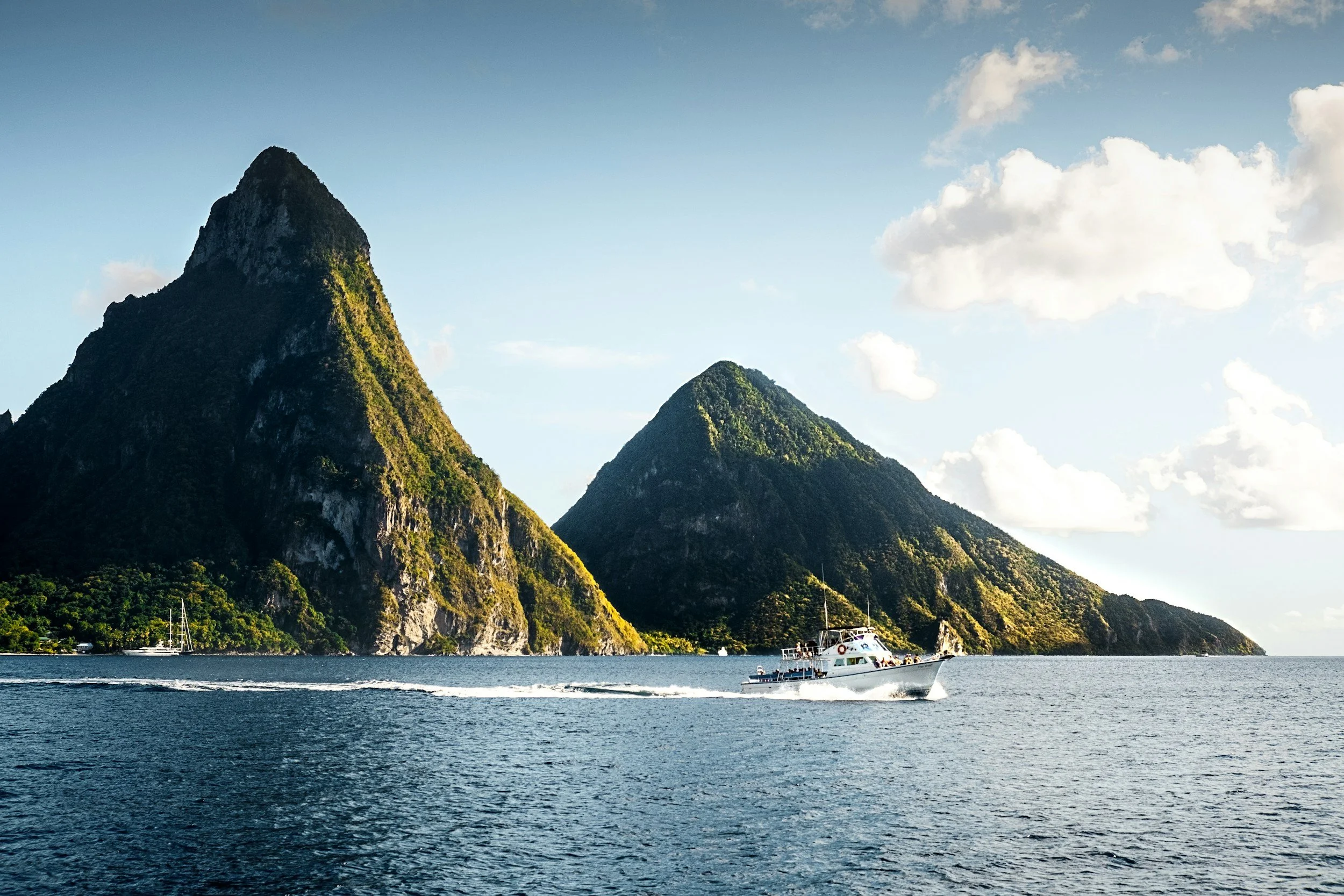 A boat cruising on the water near large green-covered mountains with a partly cloudy blue sky.