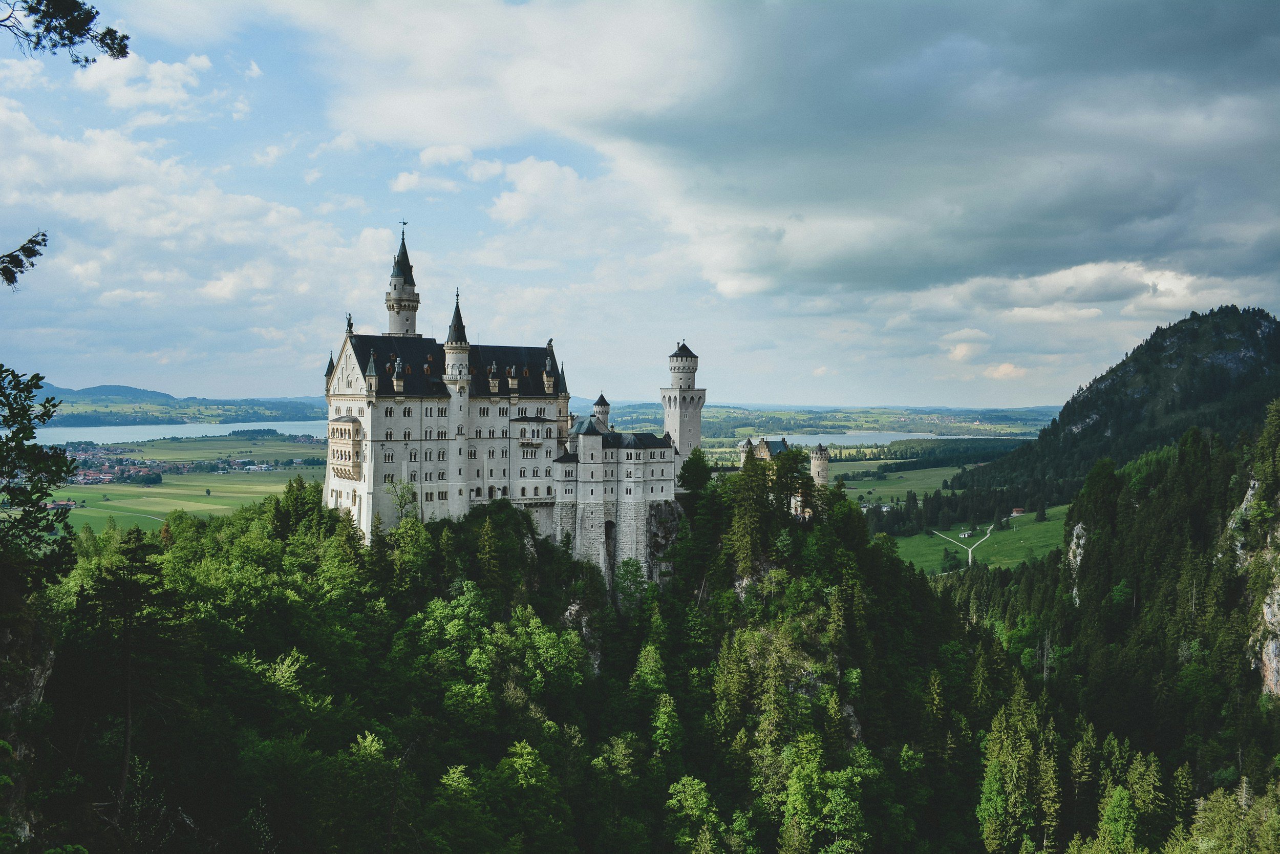 A fairy-tale castle on a green hill with surrounding trees, overlooking a scenic landscape with fields and mountains under a partly cloudy sky.