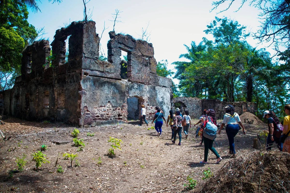 A group of people walking around the ruins of an old, abandoned brick building surrounded by trees and plants.
