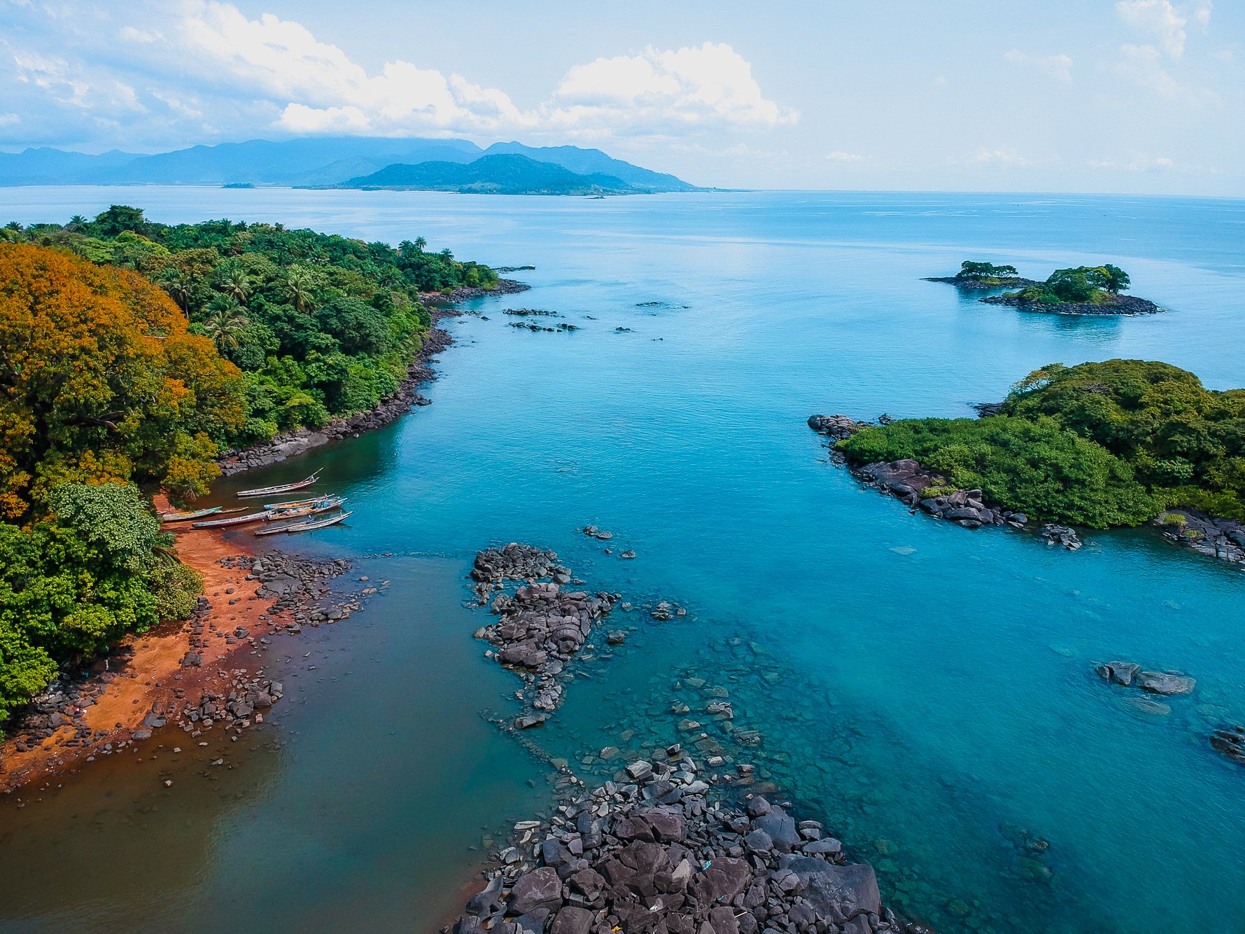 Aerial view of a tropical coastline with dense green trees, rocky shores, and clear blue water, with islands and mountains in the distance under a partly cloudy sky.