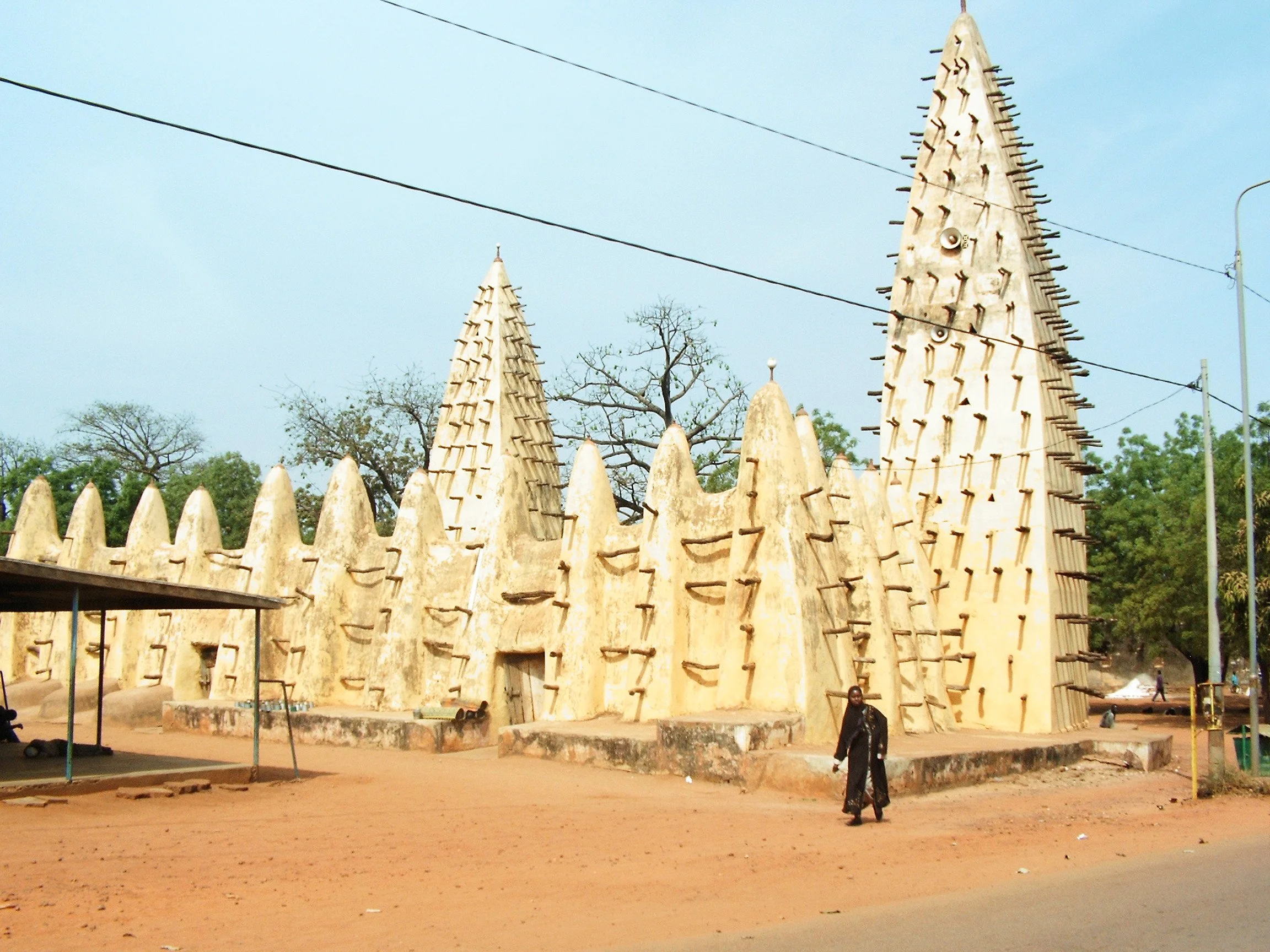 Traditional African mud building with tall, pointed towers and wooden beams, located outdoors with trees and a person walking nearby.