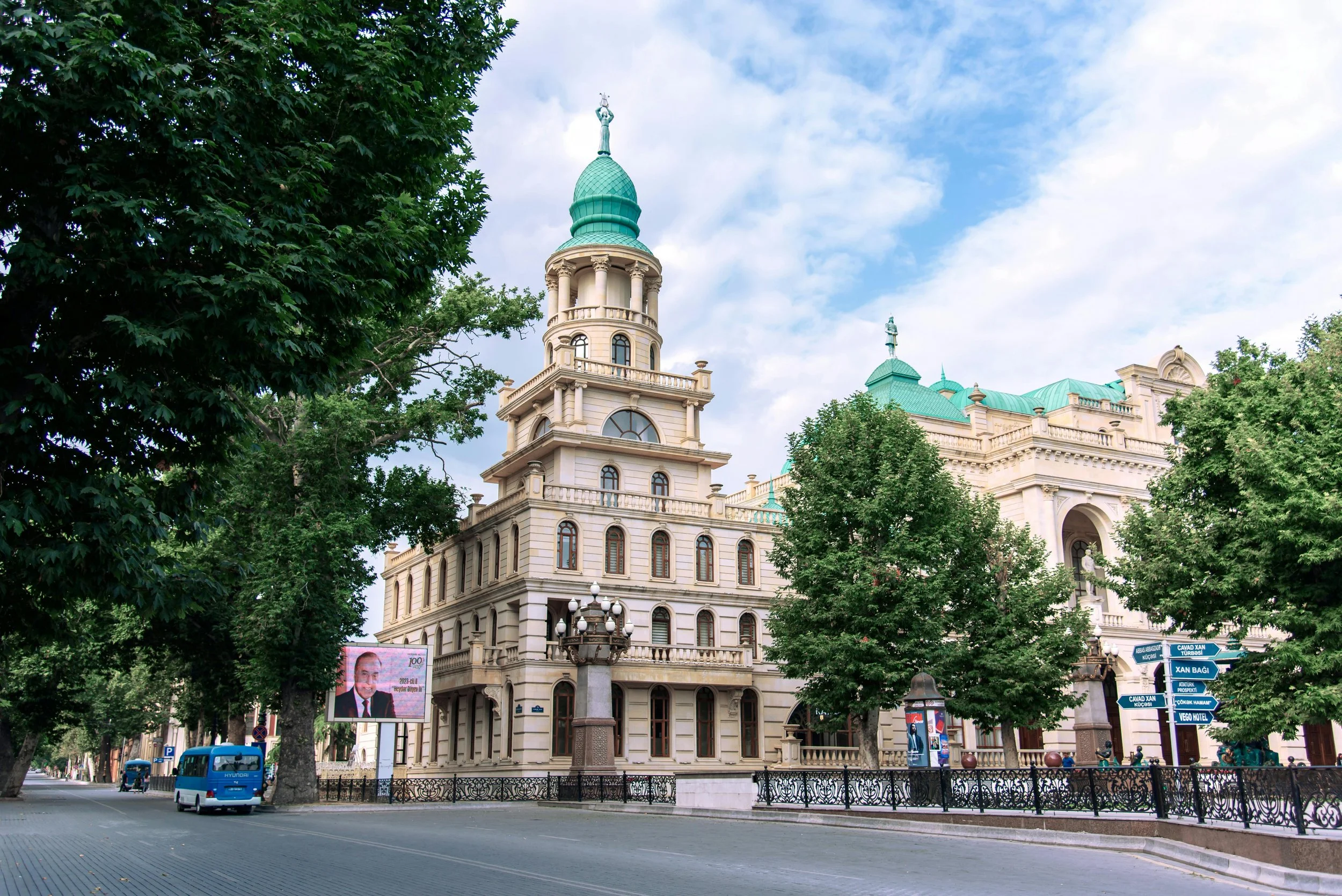A historic building with cream-colored walls and green domes, surrounded by trees and a street with a small bus and signs.