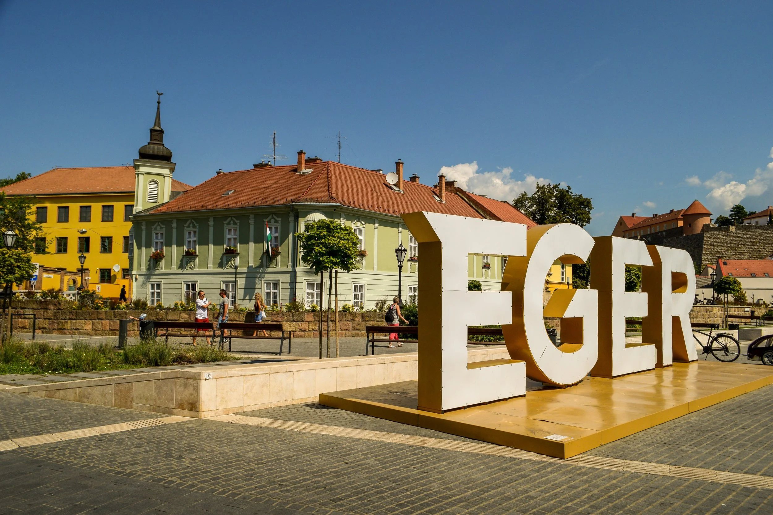 Large white and gold sign spelling 'EGER' in front of colorful buildings in a town square, with people walking and trees around.