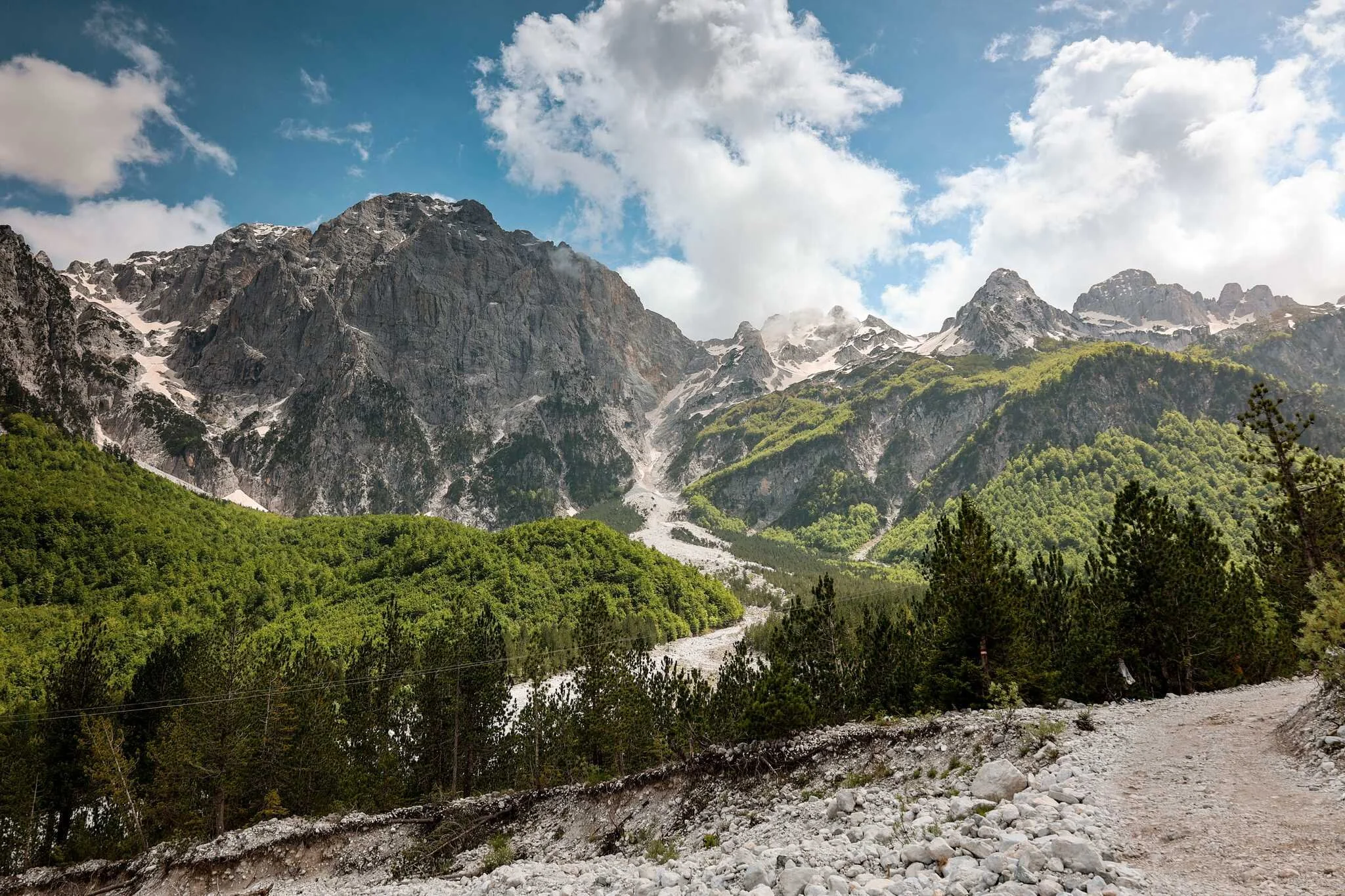 A rugged dirt trail runs along a mountain landscape with rocky terrain and sparse trees in the foreground. Behind, lush green forest covers the lower mountains, which rise to snow-capped peaks under a partly cloudy blue sky.